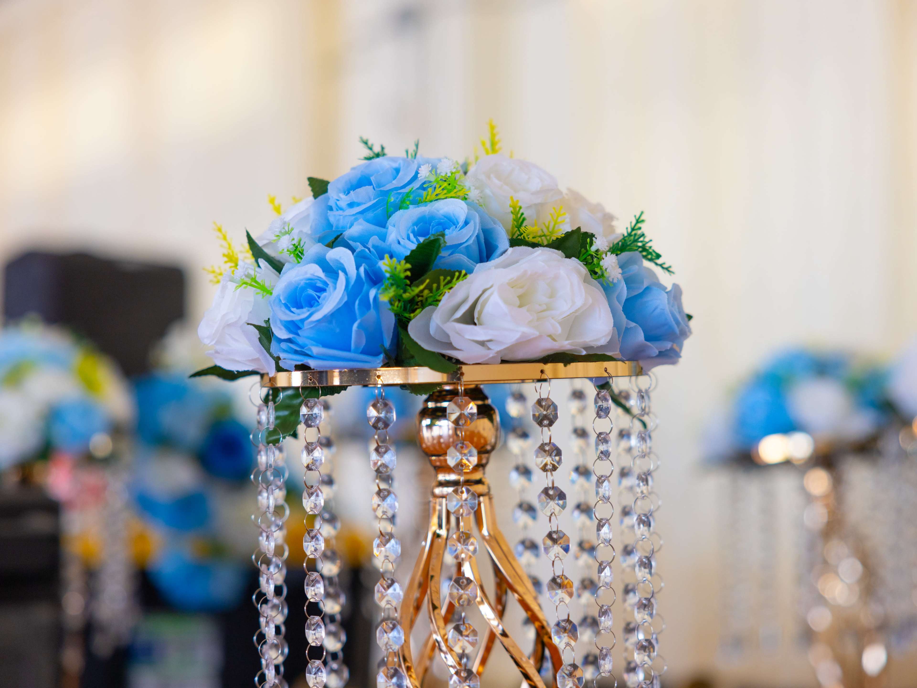 A floral arrangement of blue and white roses is displayed in a decorative gold holder adorned with hanging crystals.