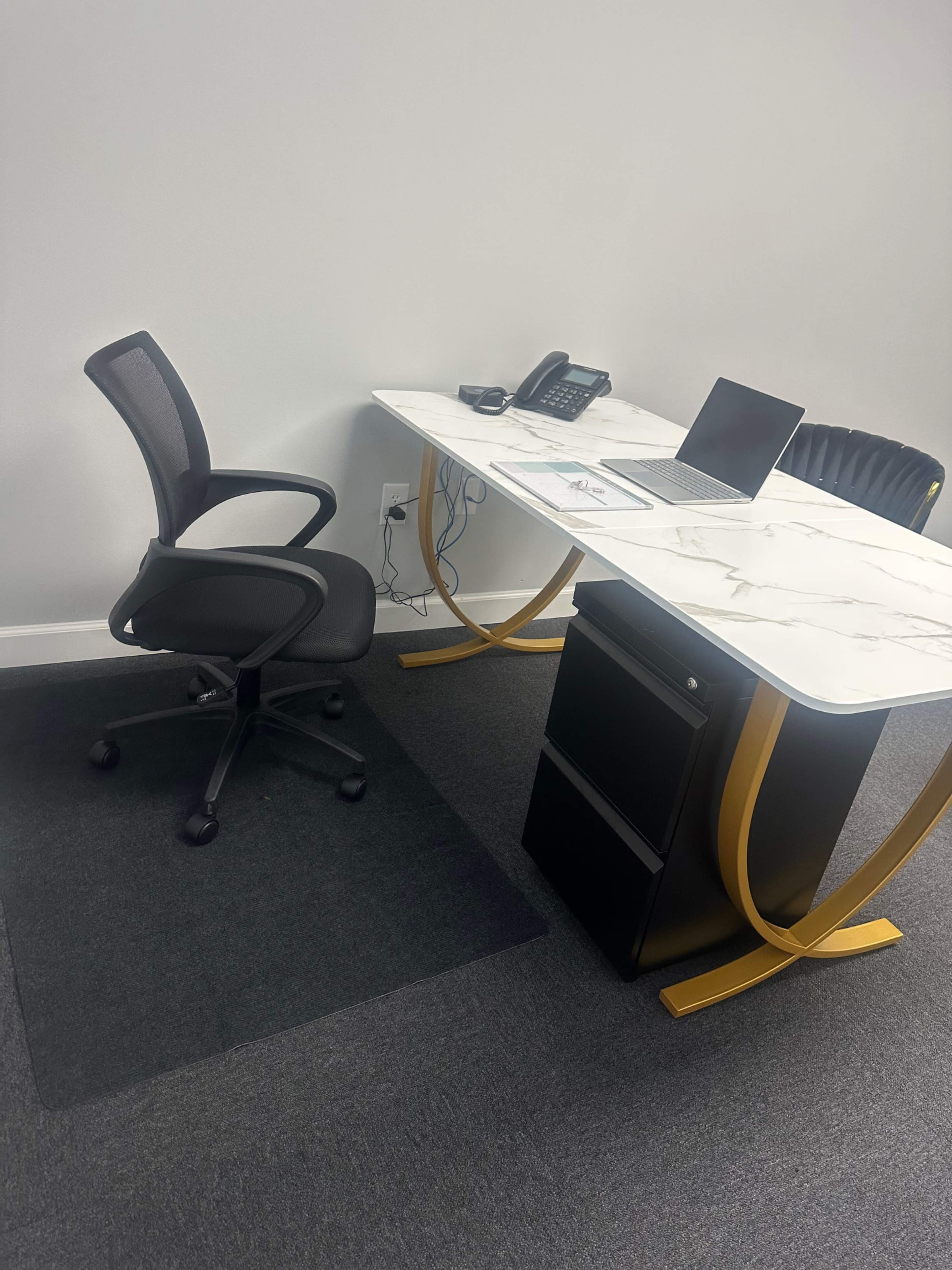 A modern office desk with a marble top, a laptop, a telephone, and a black rolling chair on a gray carpet.
