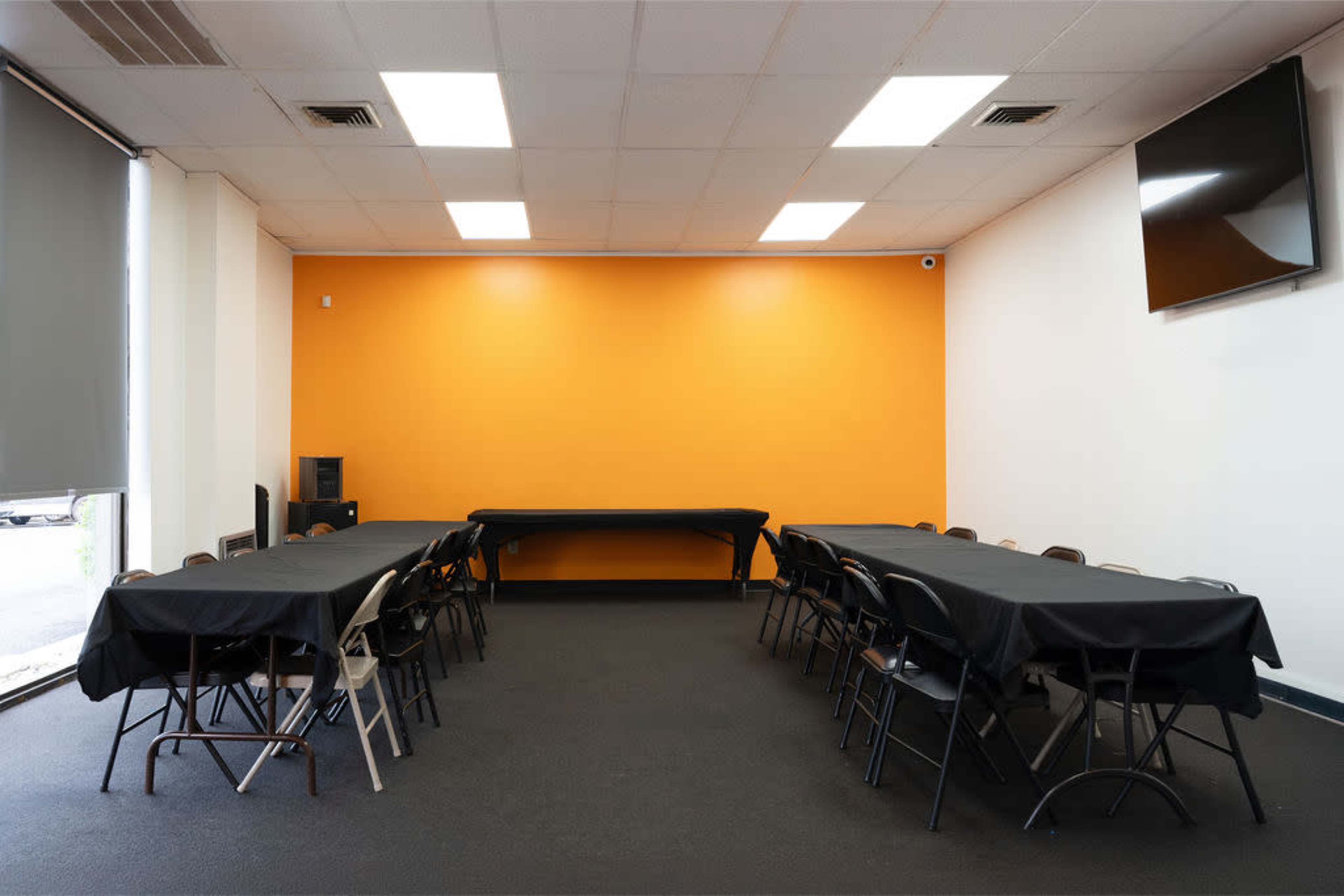 The image shows a conference room with two rows of black-covered tables arranged in a U-shape, featuring black chairs and an orange accent wall.