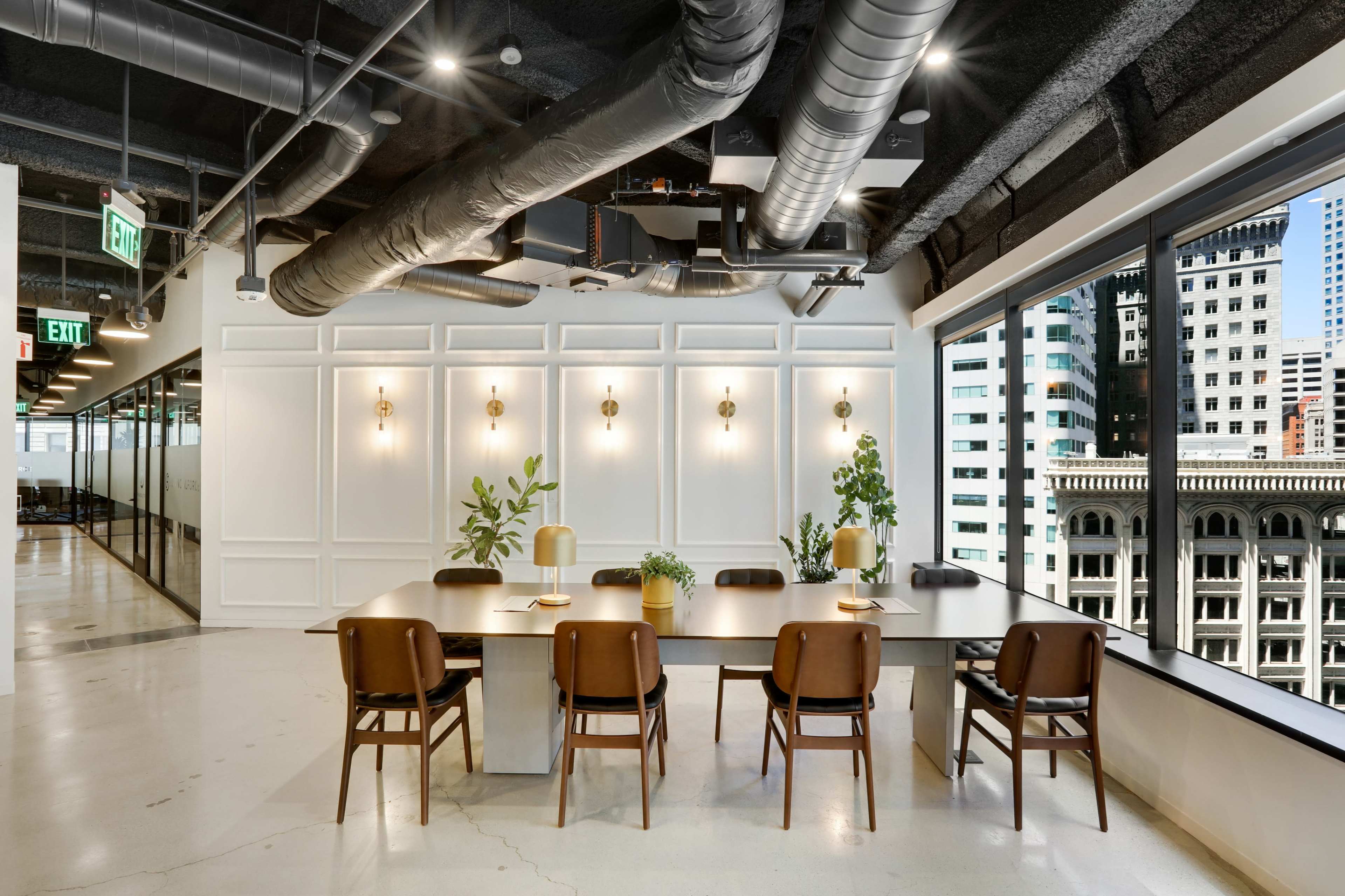 A modern conference room features a large wooden table surrounded by six chairs, with plants and lamps on the table and large windows showcasing a city view.