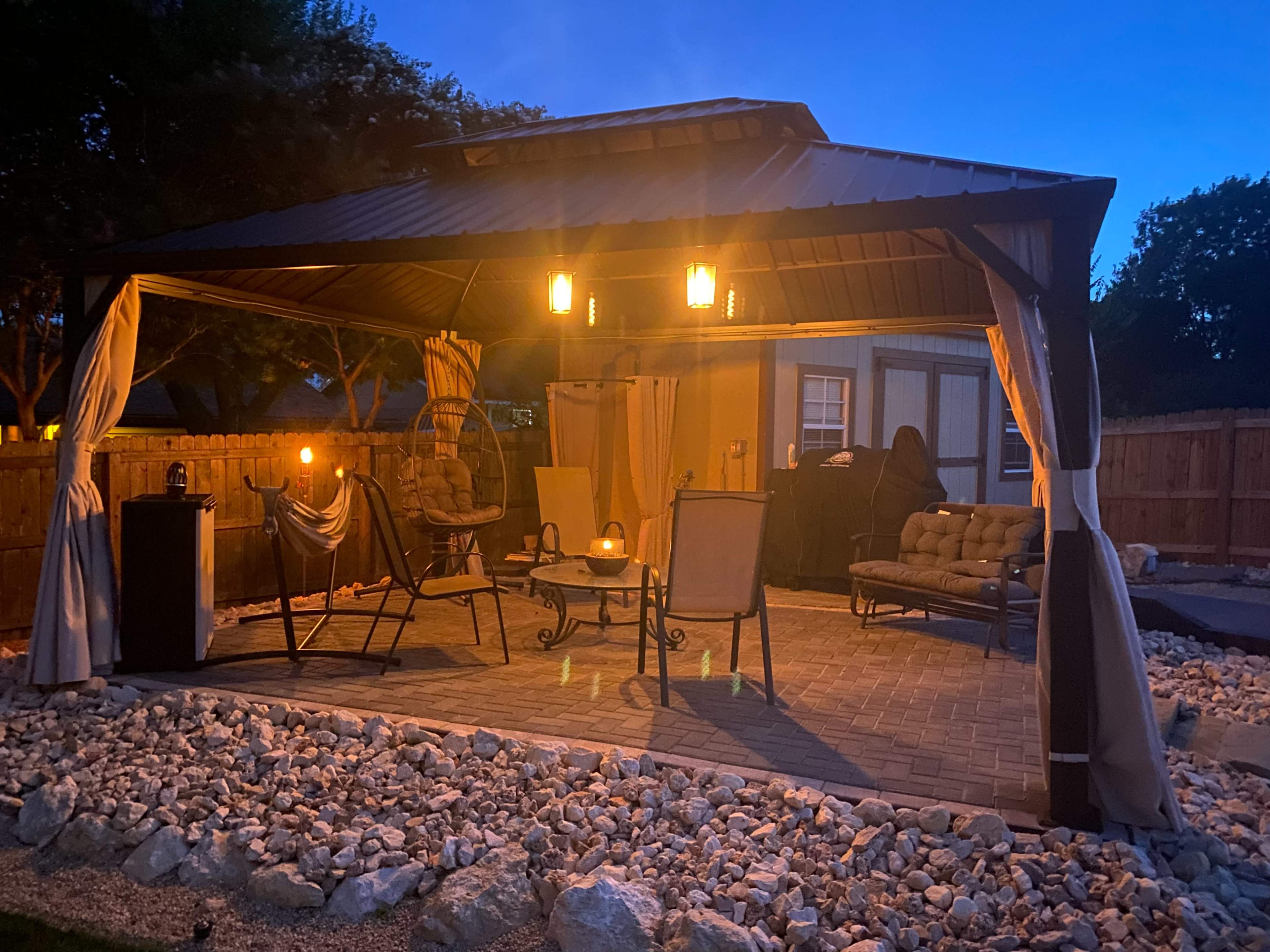 A backyard gazebo features seating, a table, and decorative stone landscaping, illuminated by pendant lights at dusk.