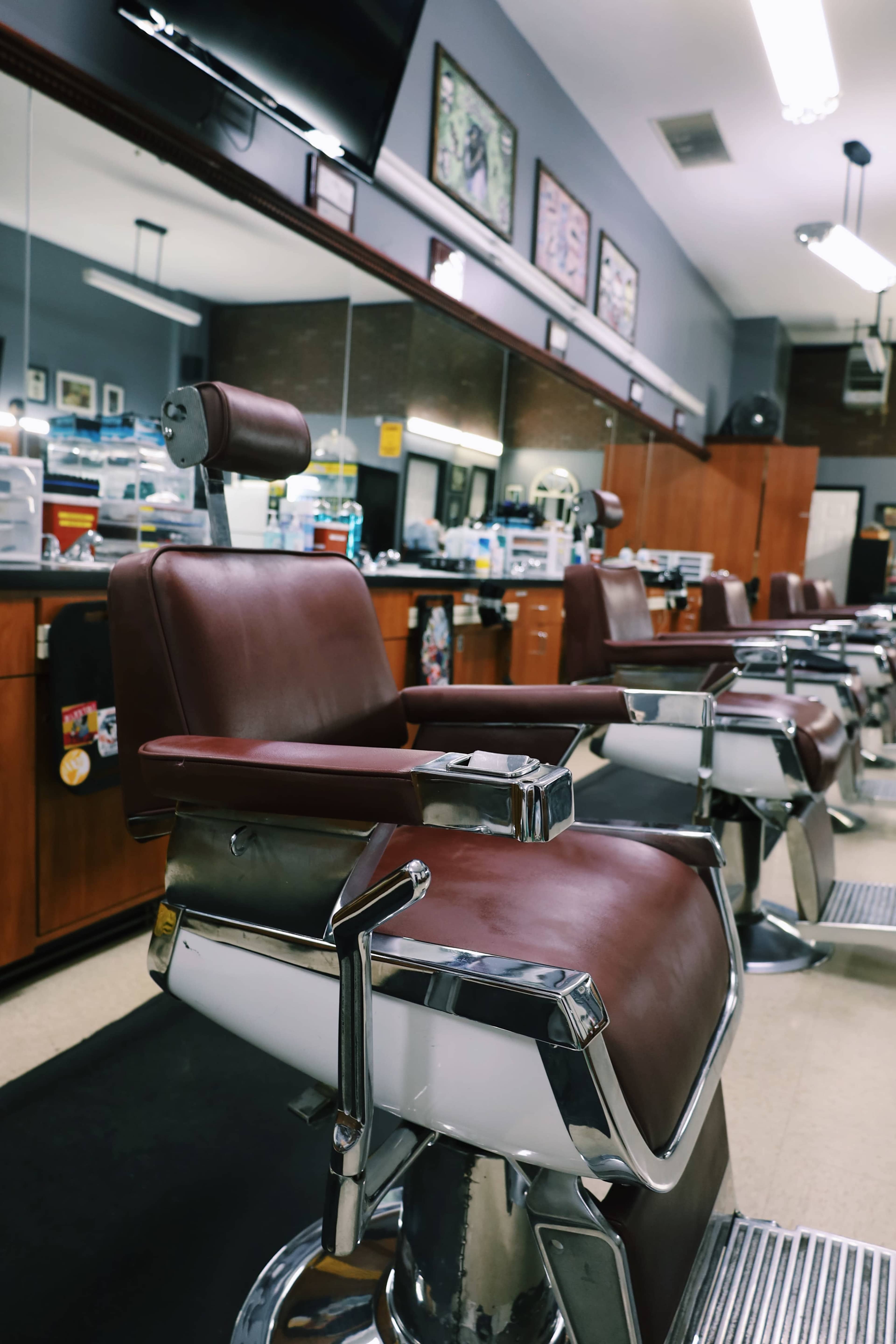 The image shows a row of brown leather barber chairs in a well-lit barbershop with mirrors and various grooming supplies in the background.