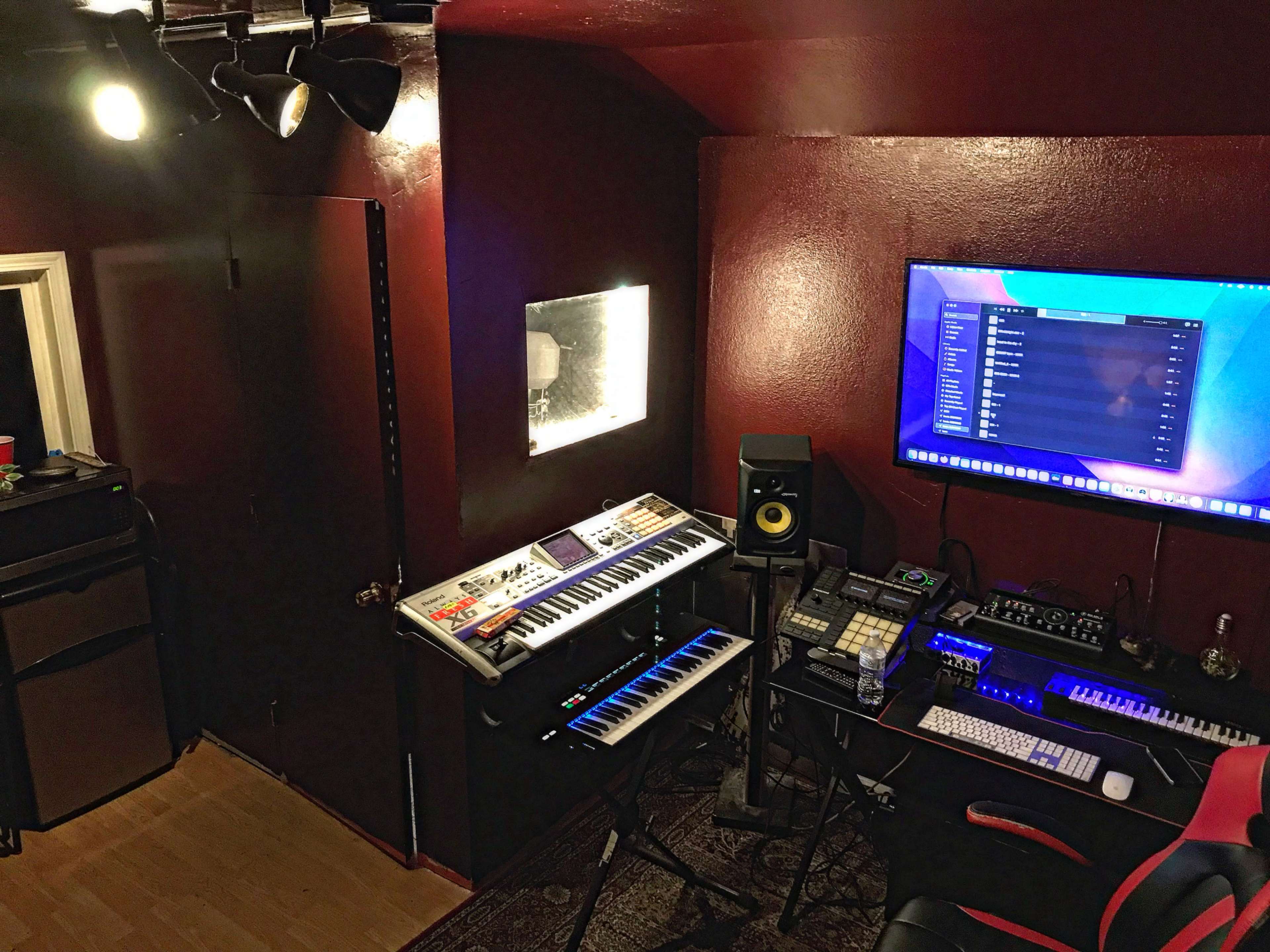 A music production studio with a computer, various synthesizers, and studio equipment against a dark red wall.