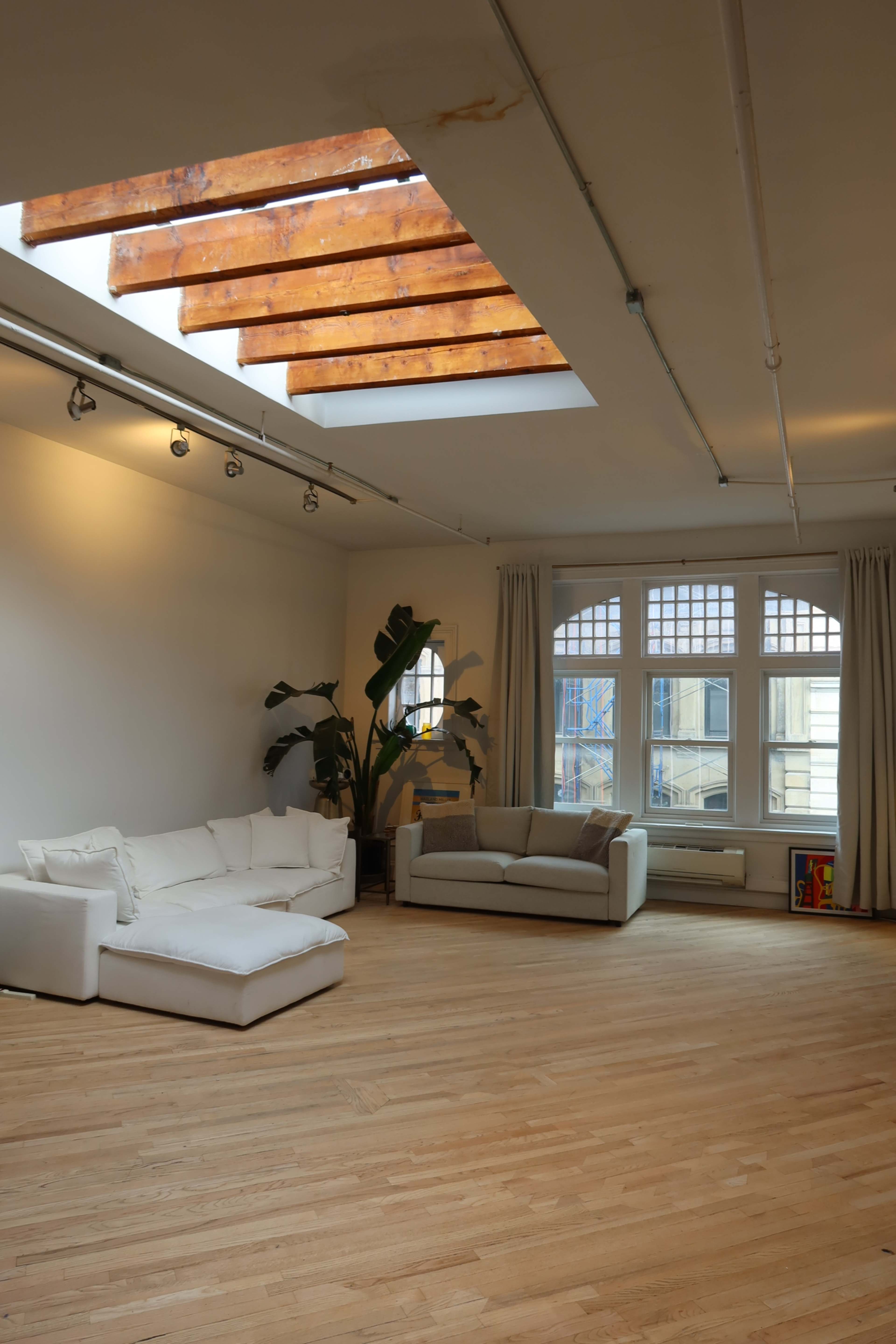 The spacious room features two light-colored sofas, large windows with wooden beams above, and a polished wooden floor.