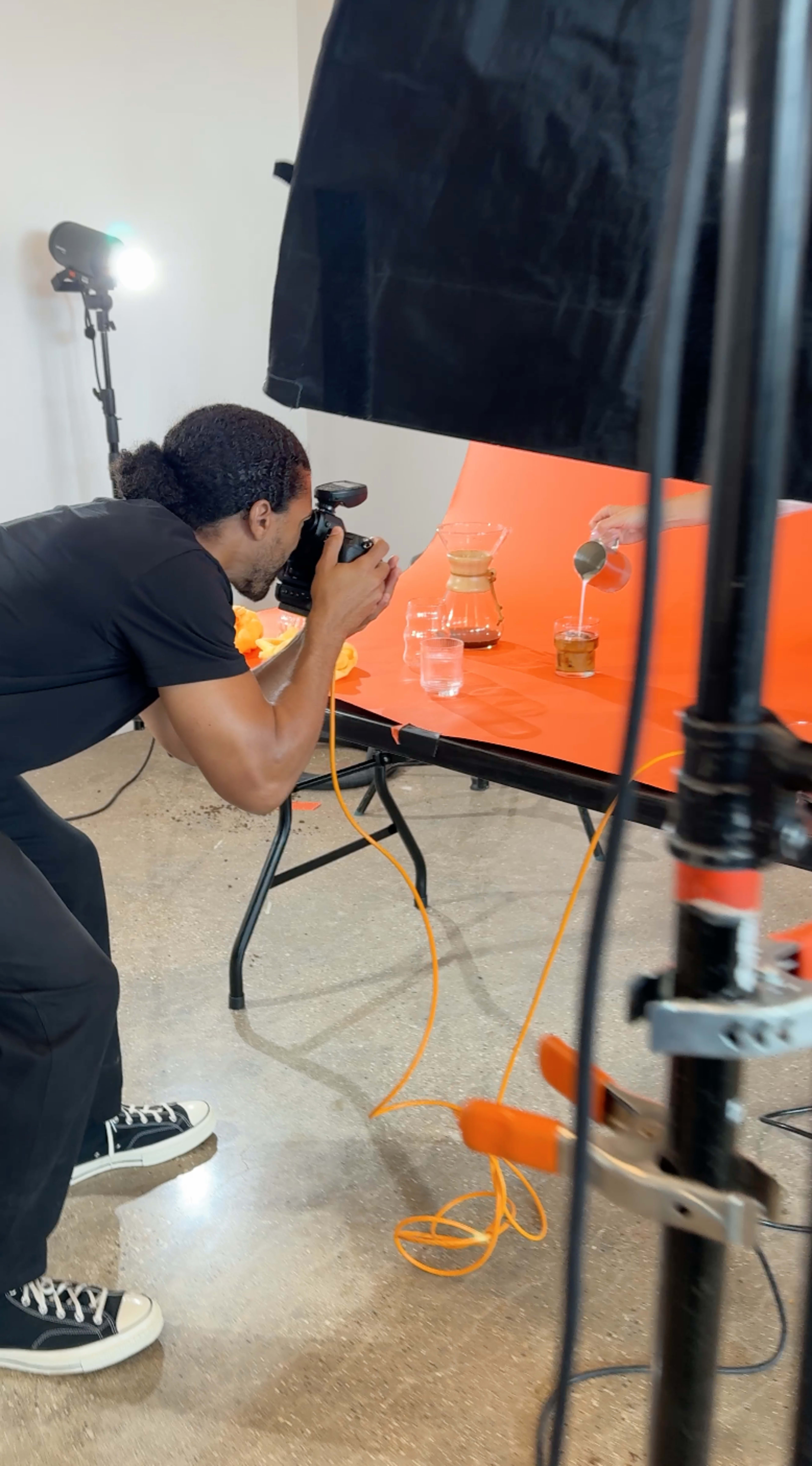 A photographer is capturing an image of a glass pitcher and a glass on an orange table, with lighting equipment positioned around the scene.