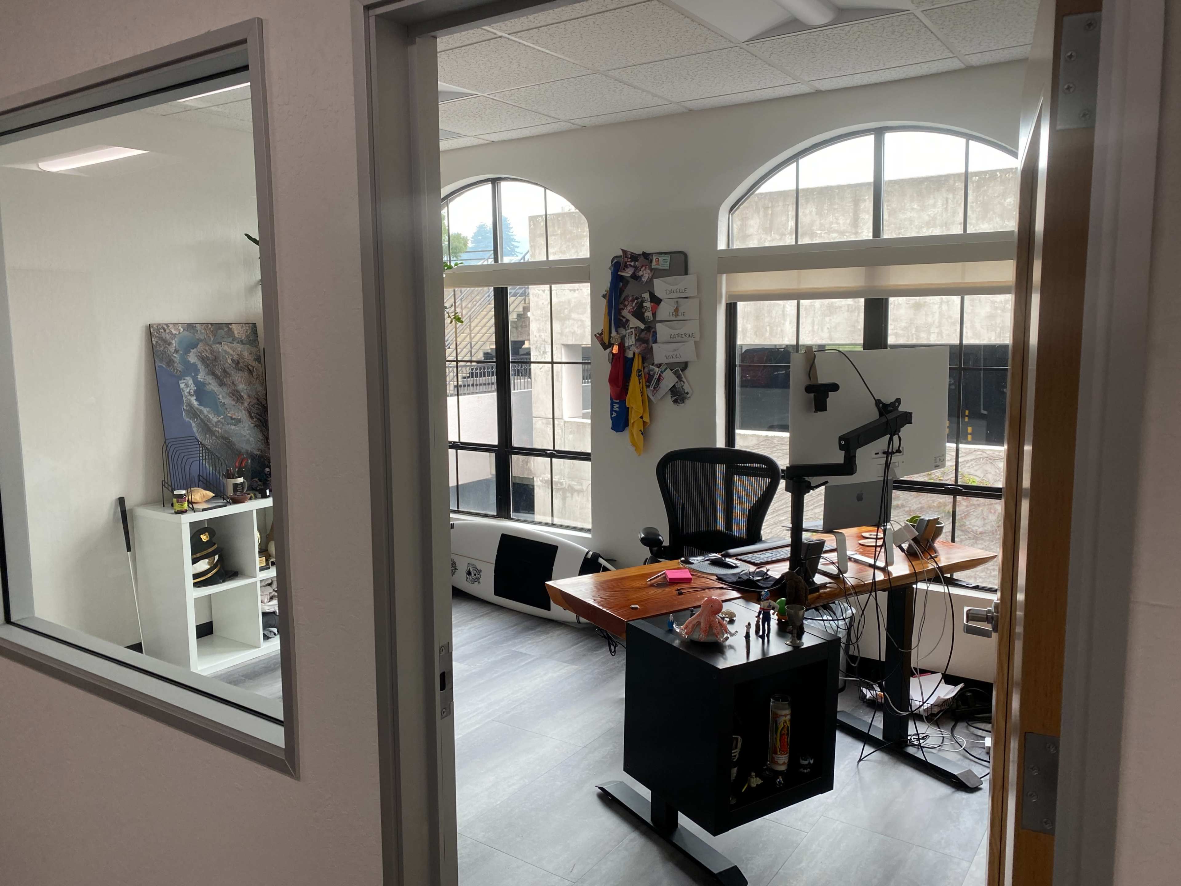 An office space with a desk, ergonomic chair, and various items on shelves, visible through a doorway.
