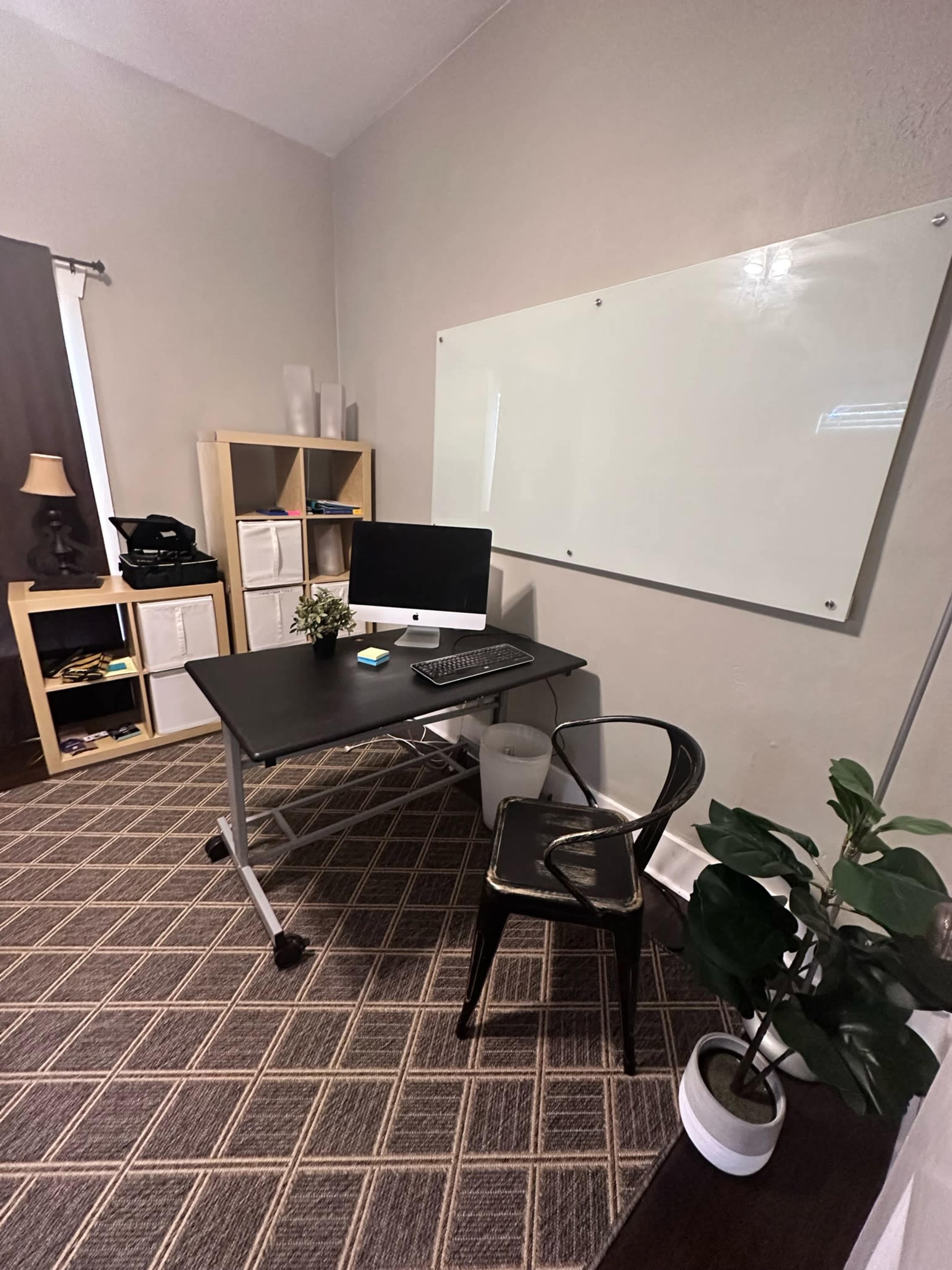 A minimalist office with a black desk, a computer setup, a chair, a whiteboard on the wall, and a few decorative plants.