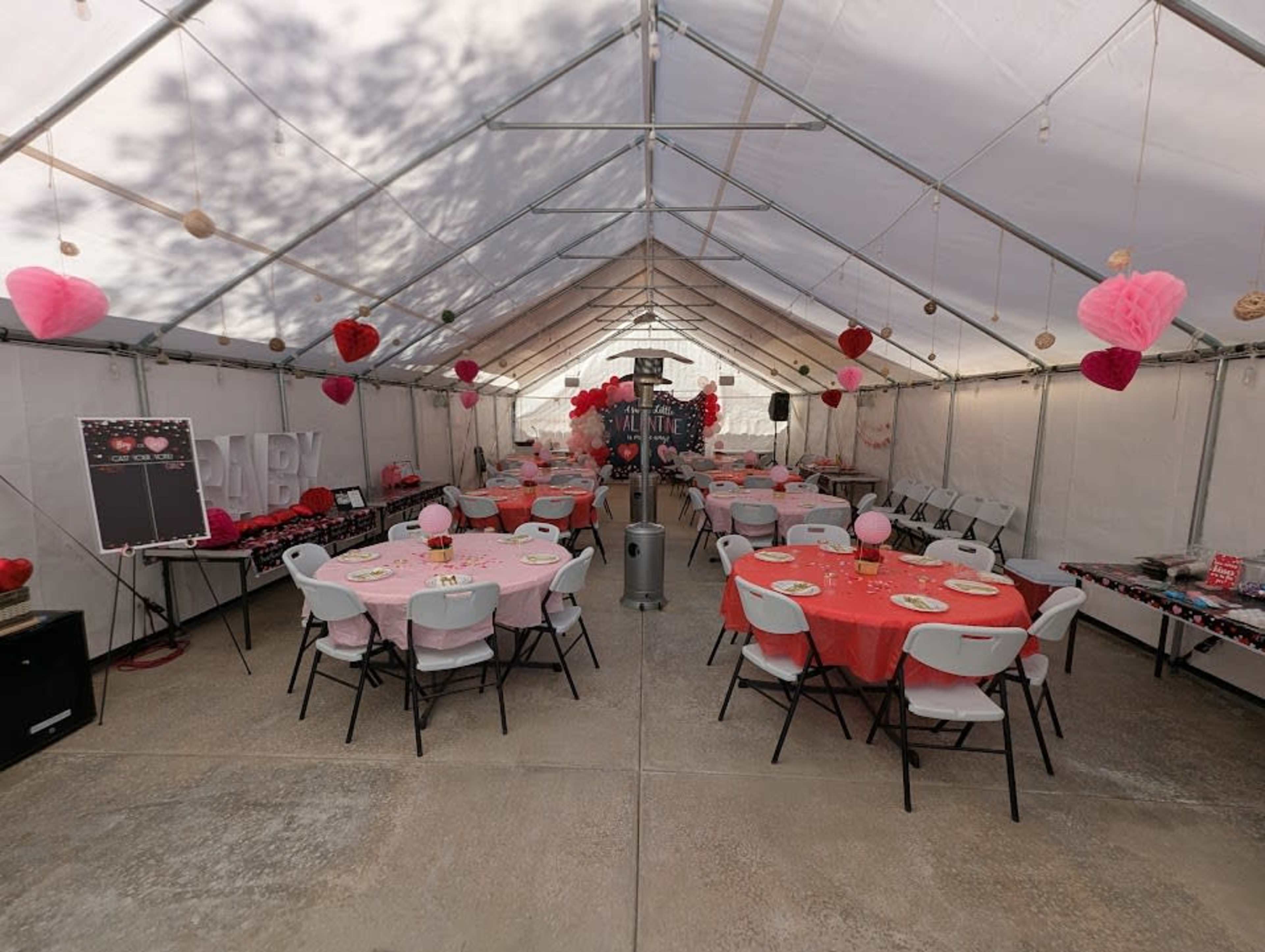 A large tent is set up for an event, featuring round tables covered with pink tablecloths, decorations including heart-shaped ornaments, and chairs arranged for guests.