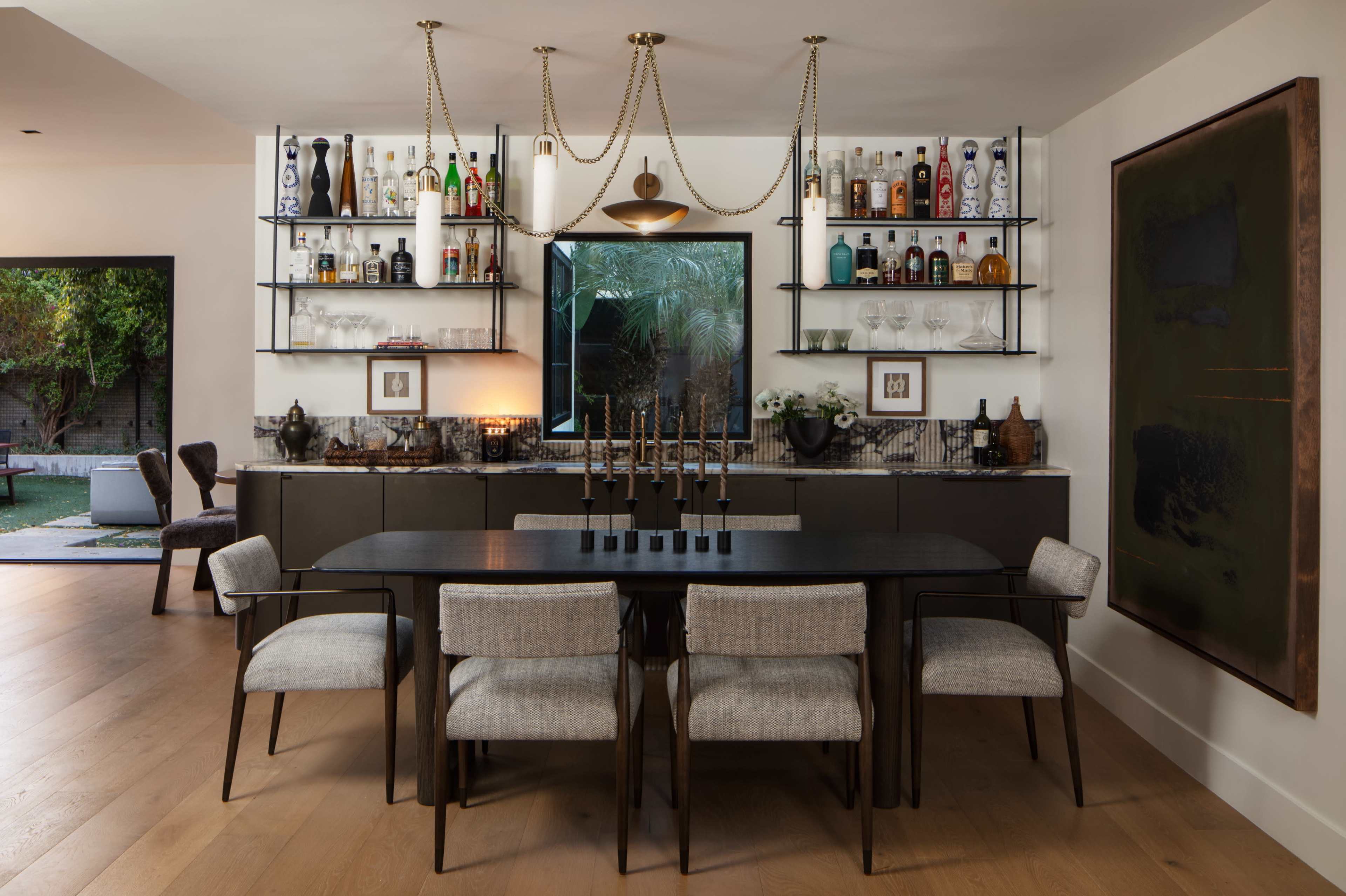 A modern dining area features a long table with upholstered chairs, a bar with various bottles on shelves, and a large mirror above, set against a backdrop of greenery.