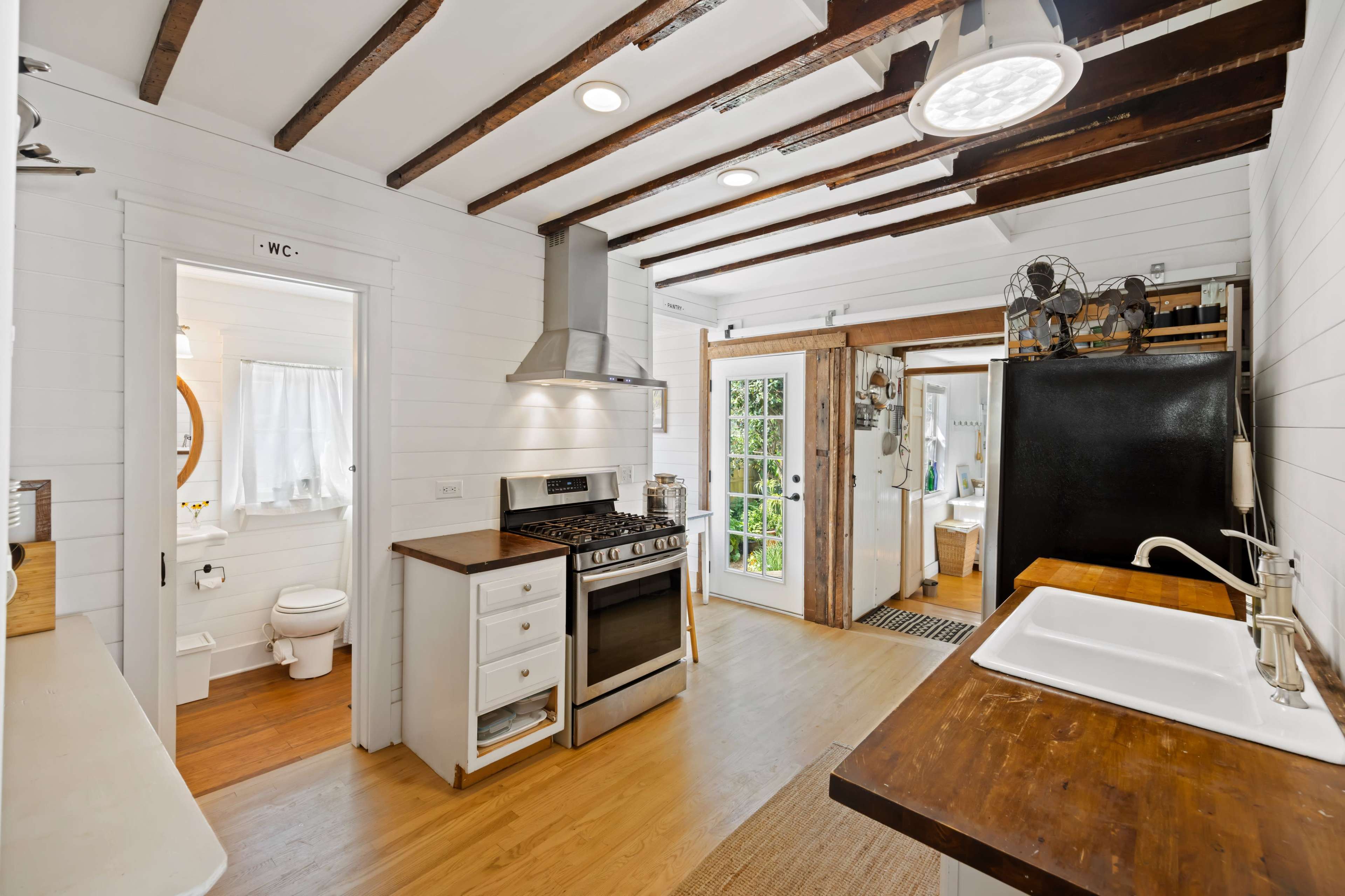 A modern kitchen features white shiplap walls, exposed wooden beams, stainless steel appliances, and a door leading to an outdoor area.