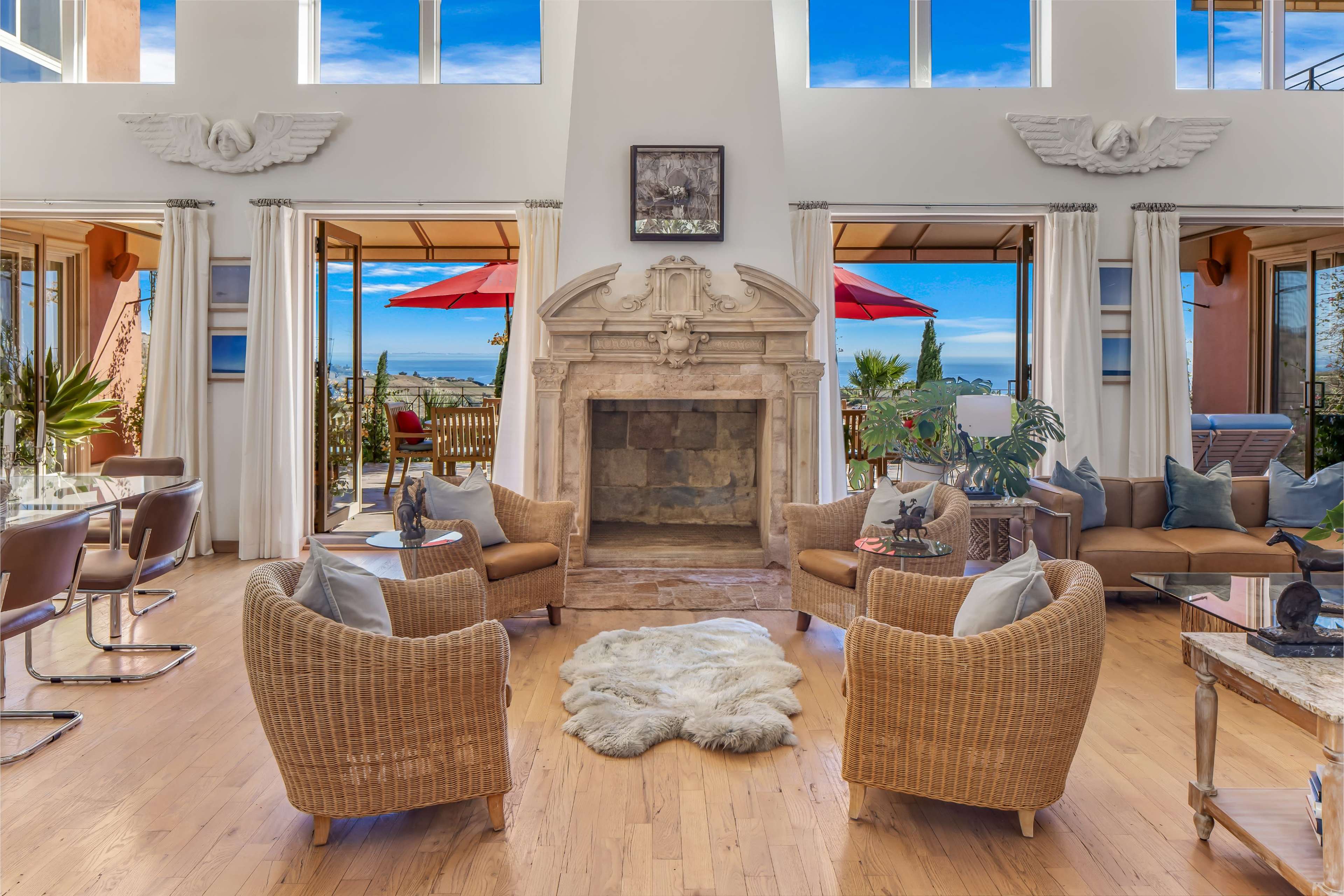 A spacious living room featuring a stone fireplace, two wicker armchairs, and large windows that offer a view of an outdoor patio with red umbrellas.
