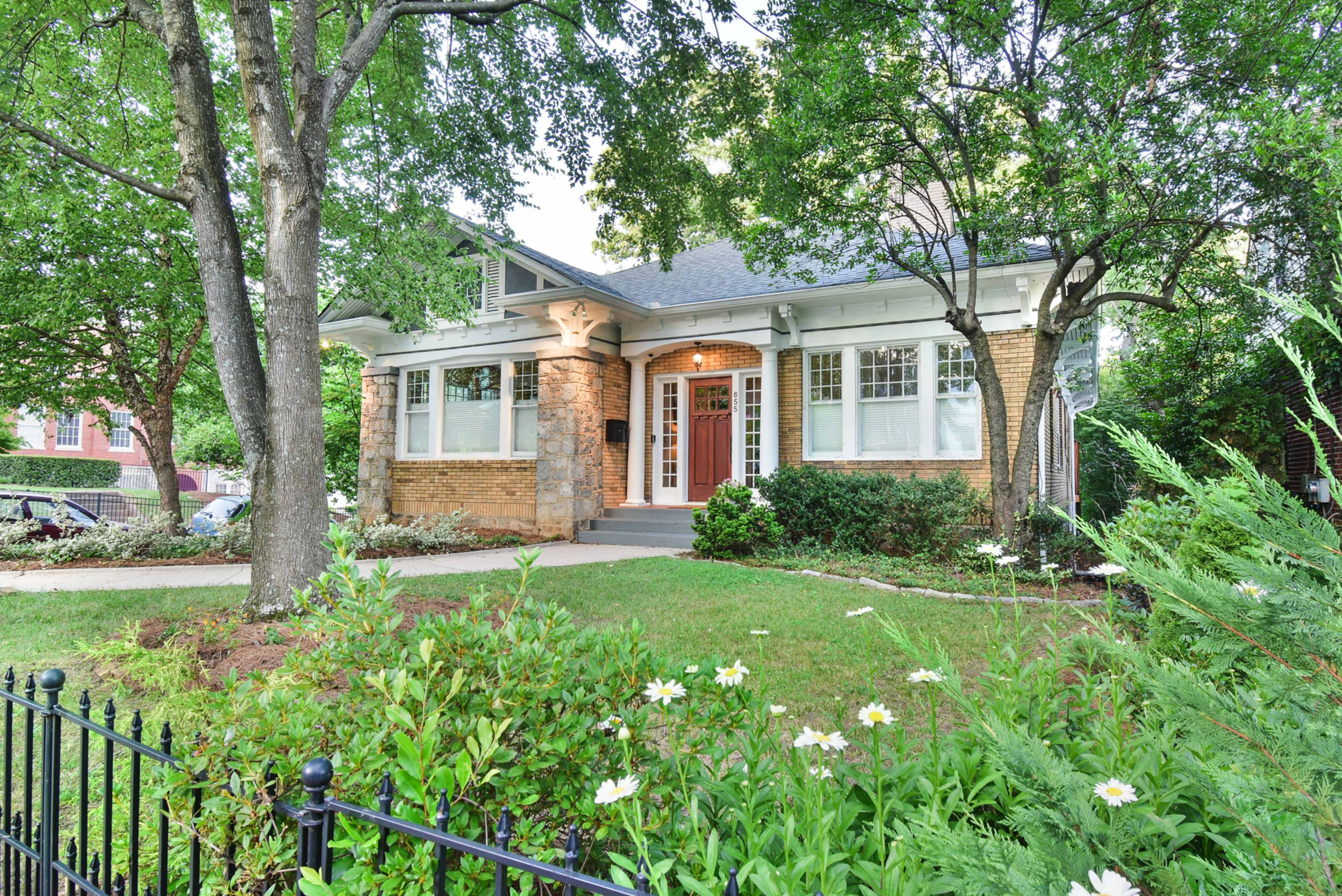 A brick house with a front porch and large windows is surrounded by greenery and flowers, with a black metal fence in the foreground.