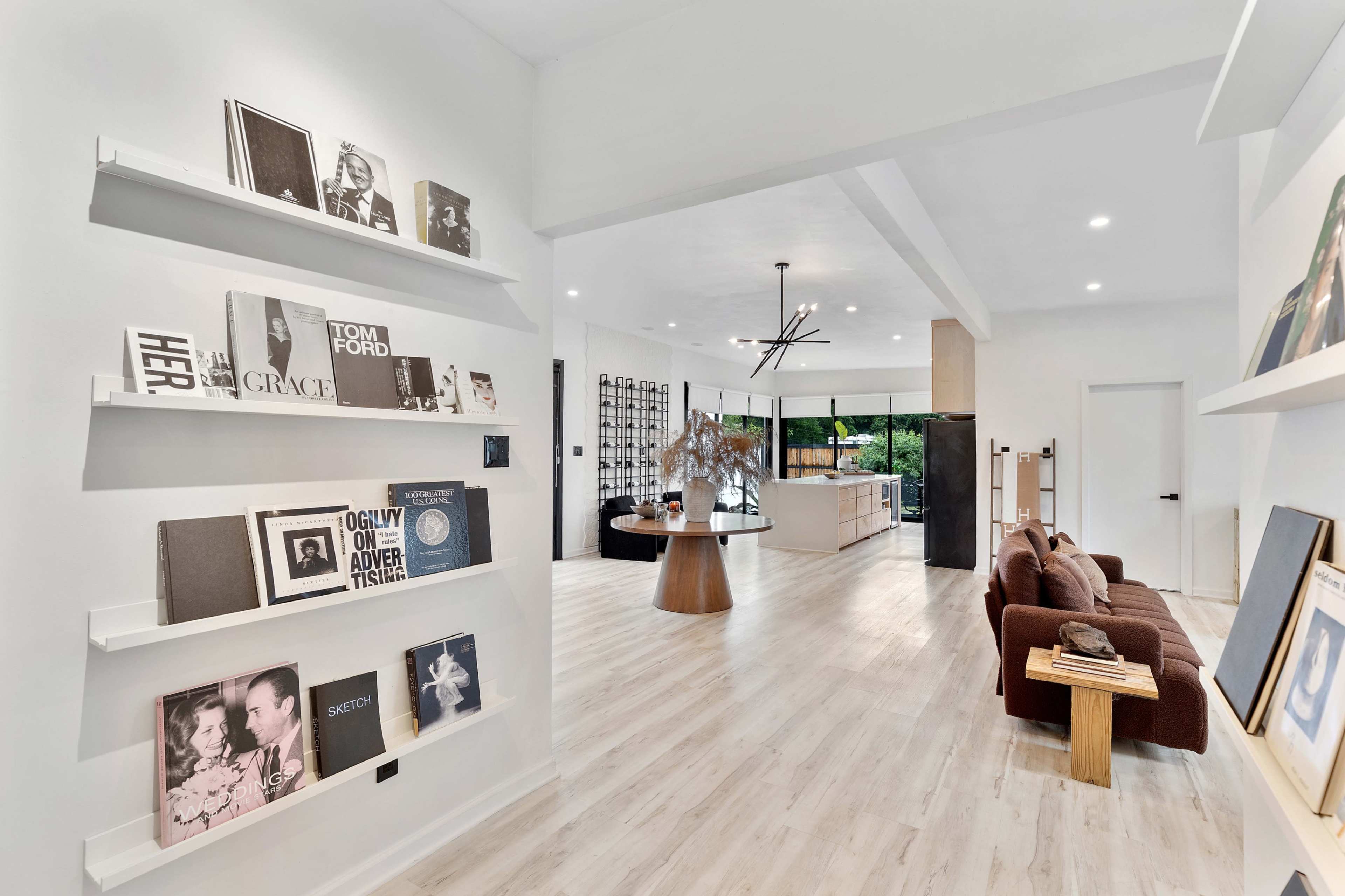 The image shows a modern interior space featuring bookshelves with various books, a cozy seating area, and a view of a kitchen and dining space in the background.