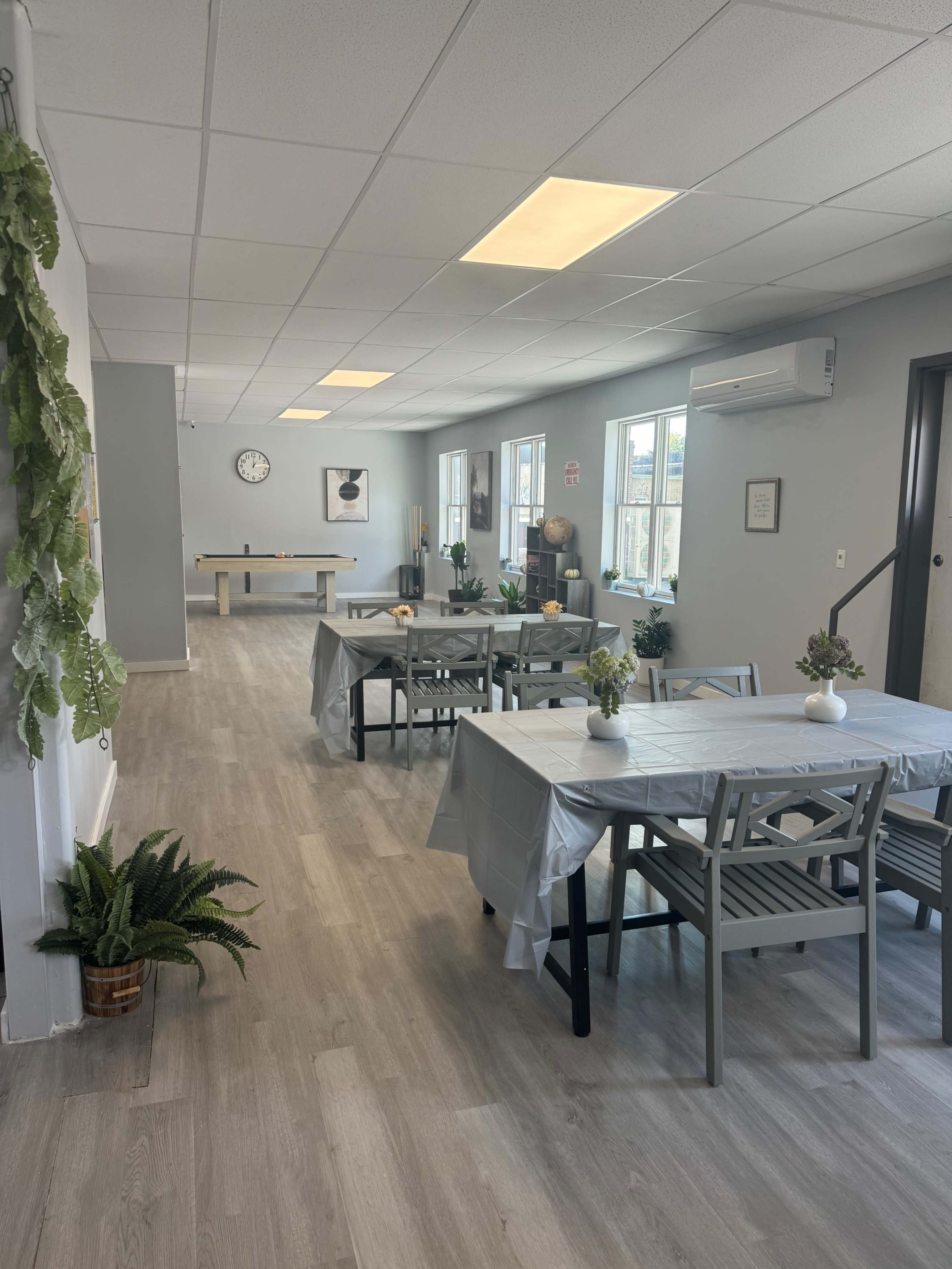 The image shows a spacious, well-lit interior with several tables set for dining, plants in pots, and a game area in the background.