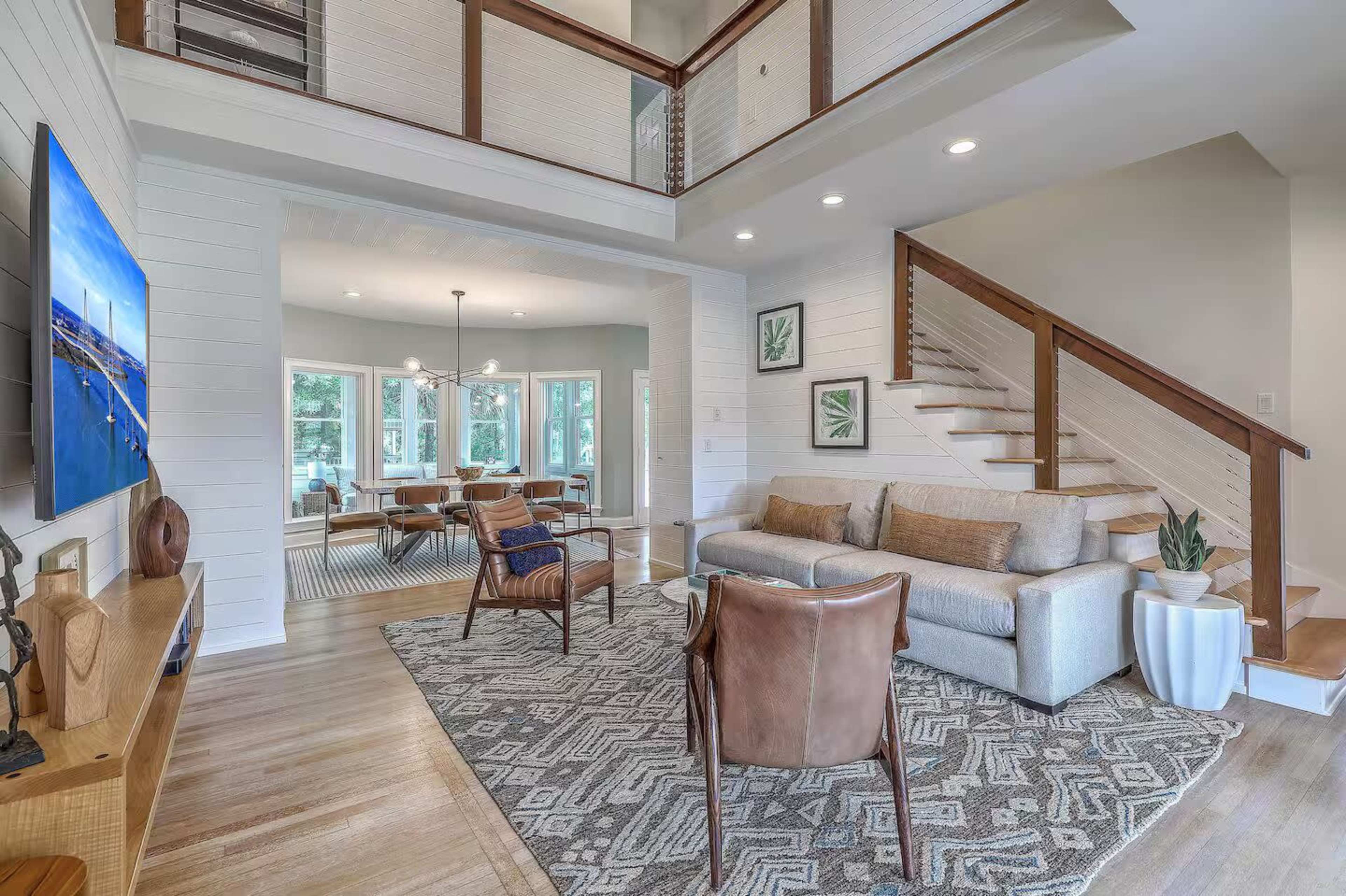 The image shows a modern living room with a comfortable sofa, accent chairs, and a staircase leading to a second level, all set against a backdrop of large windows.