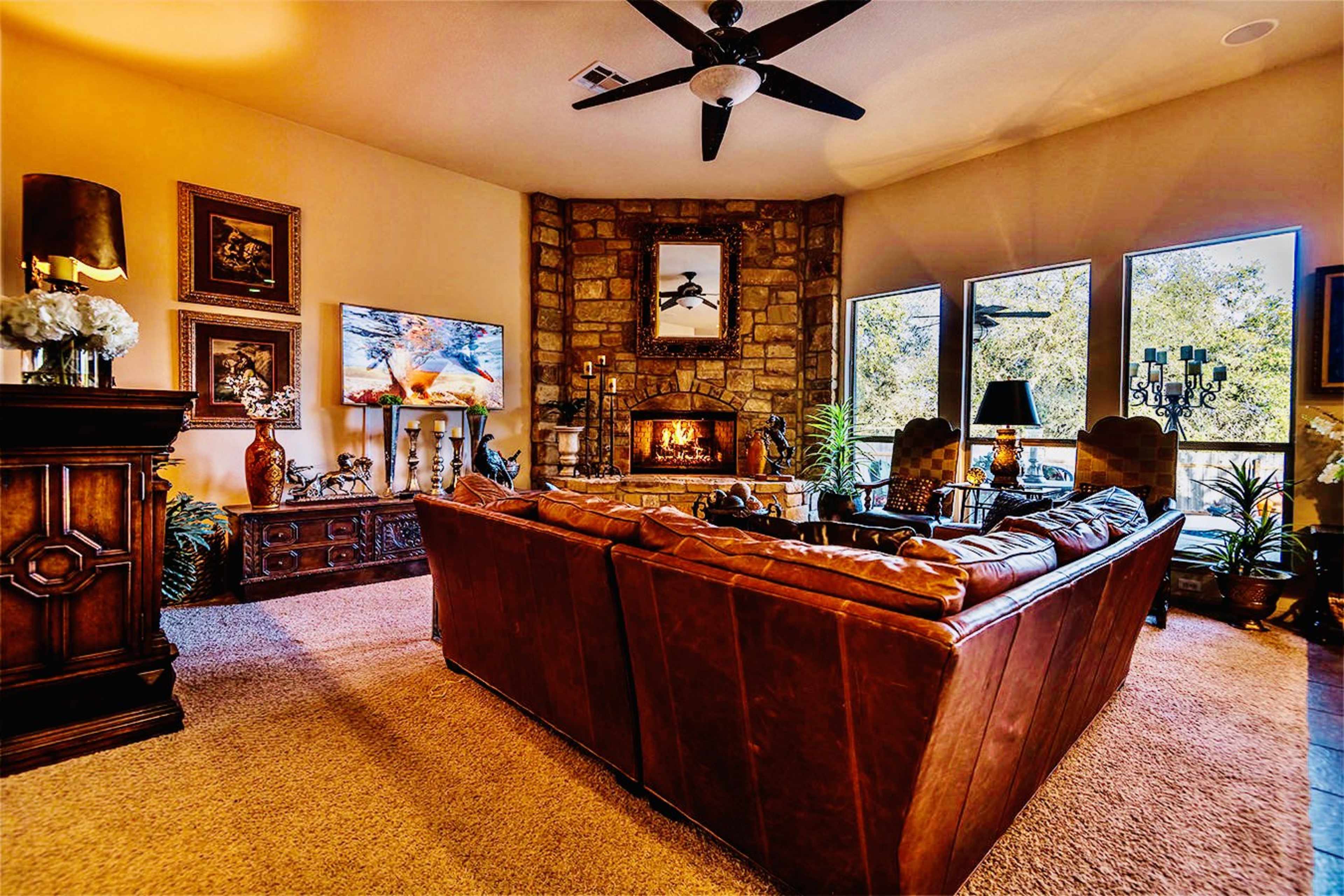 The cozy living room features a stone fireplace, large windows, and a brown leather sectional sofa arranged around a central coffee table.