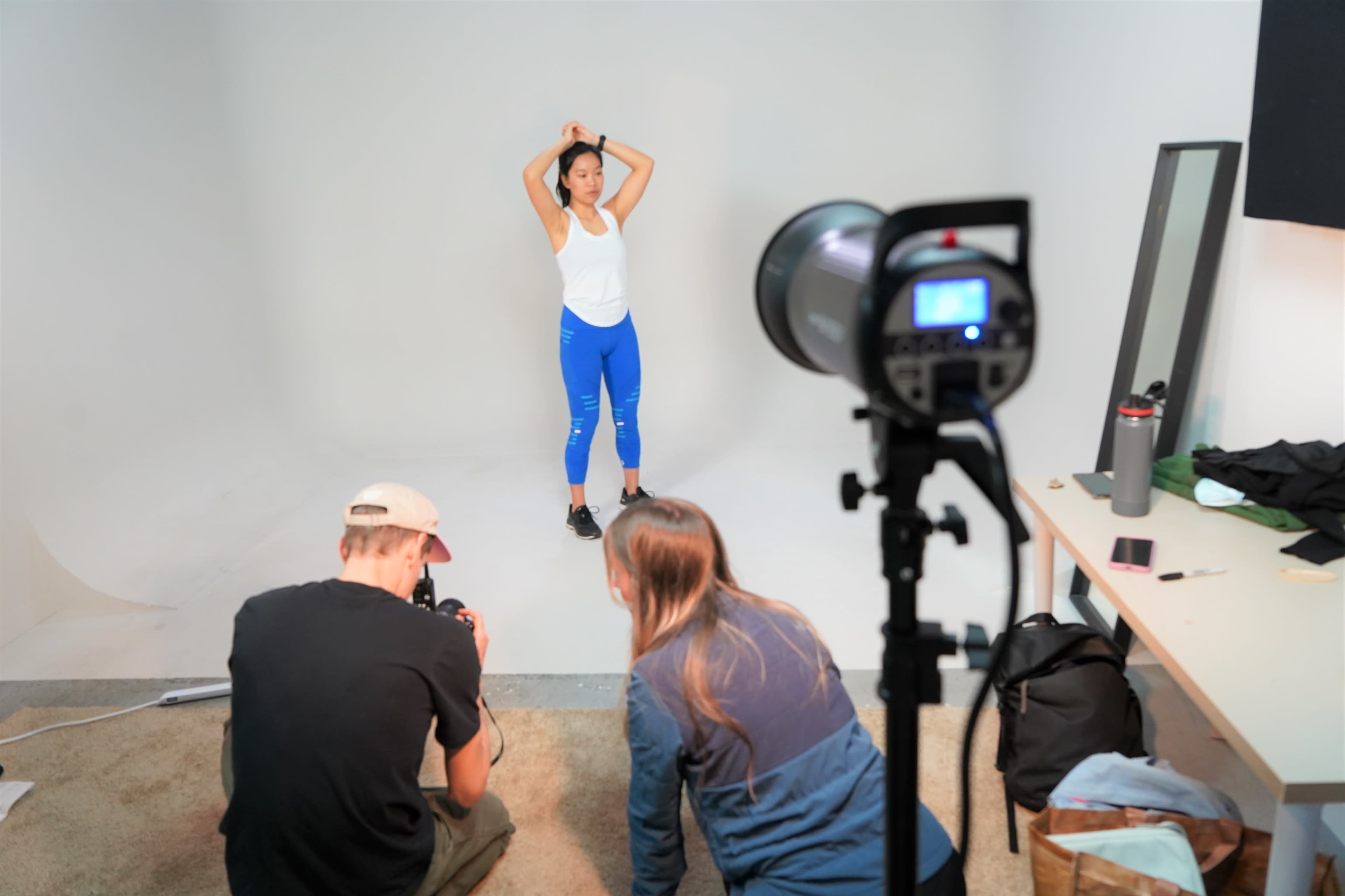 A model poses in a studio while two photographers set up equipment and capture images.