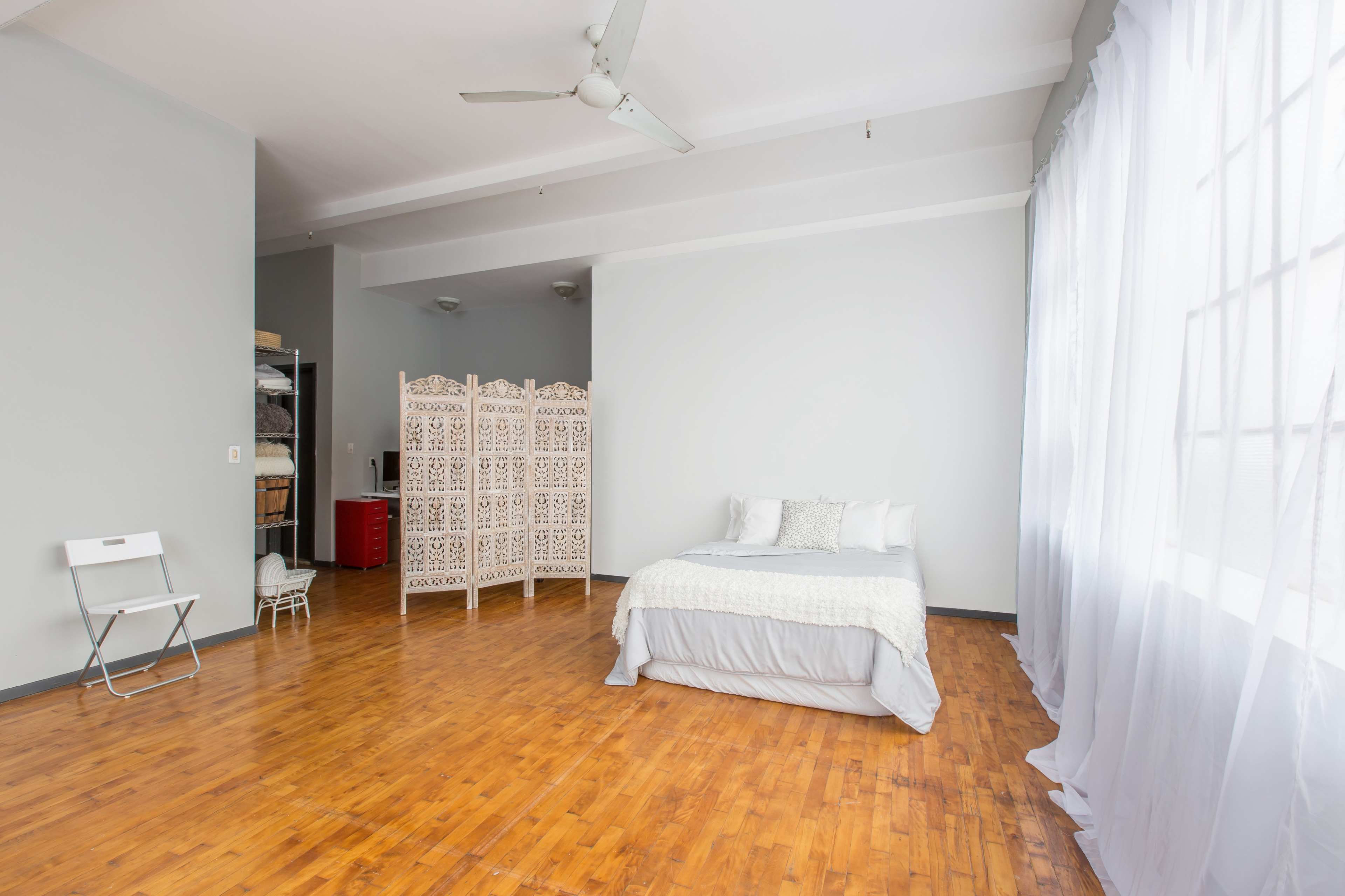 The image shows a spacious room with wooden flooring, a bed against the wall, a privacy screen in the center, and large windows covered with sheer curtains.