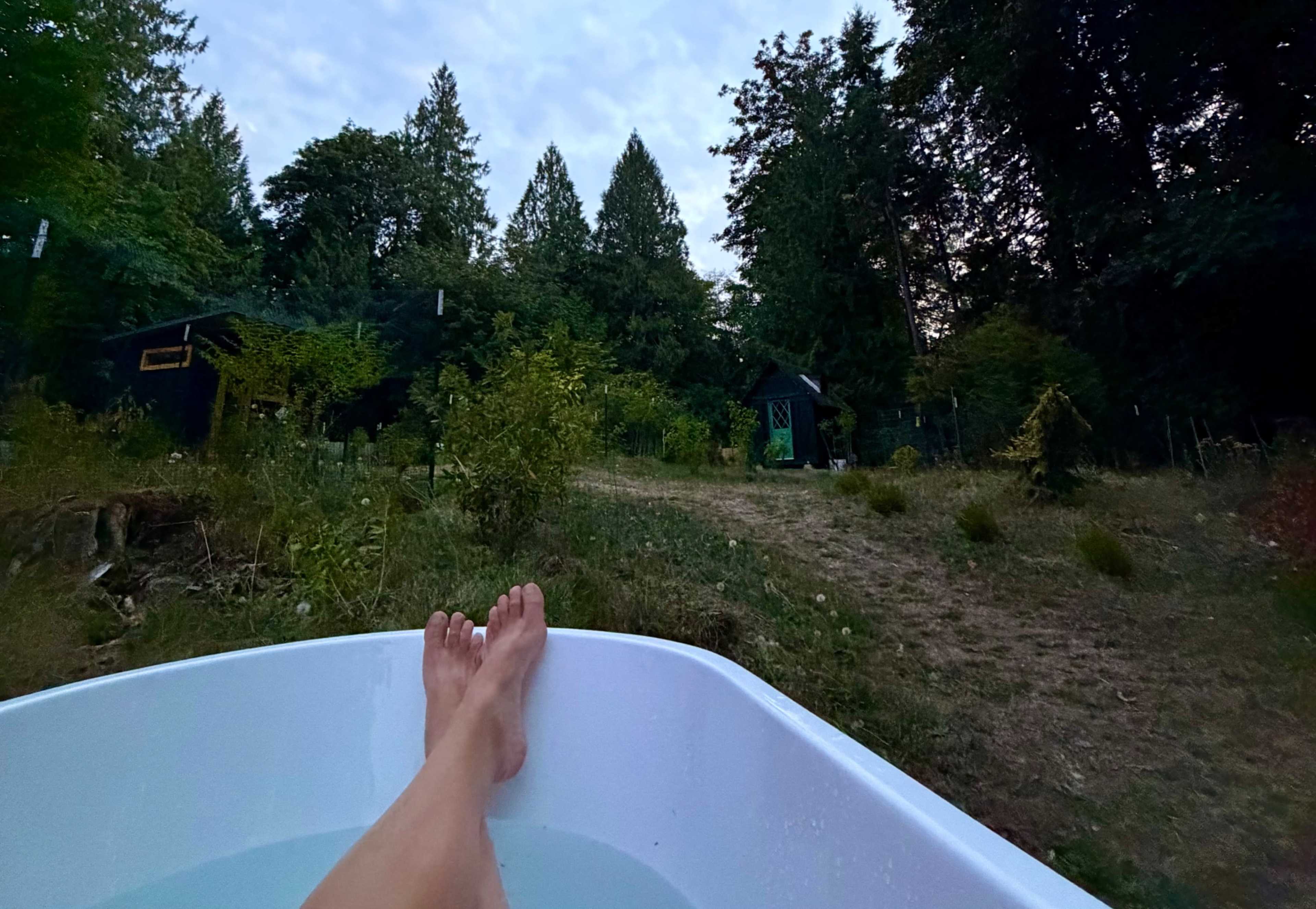 A person lounges in a white bathtub filled with water, with a view of a natural landscape featuring trees and a path leading to a small green building.