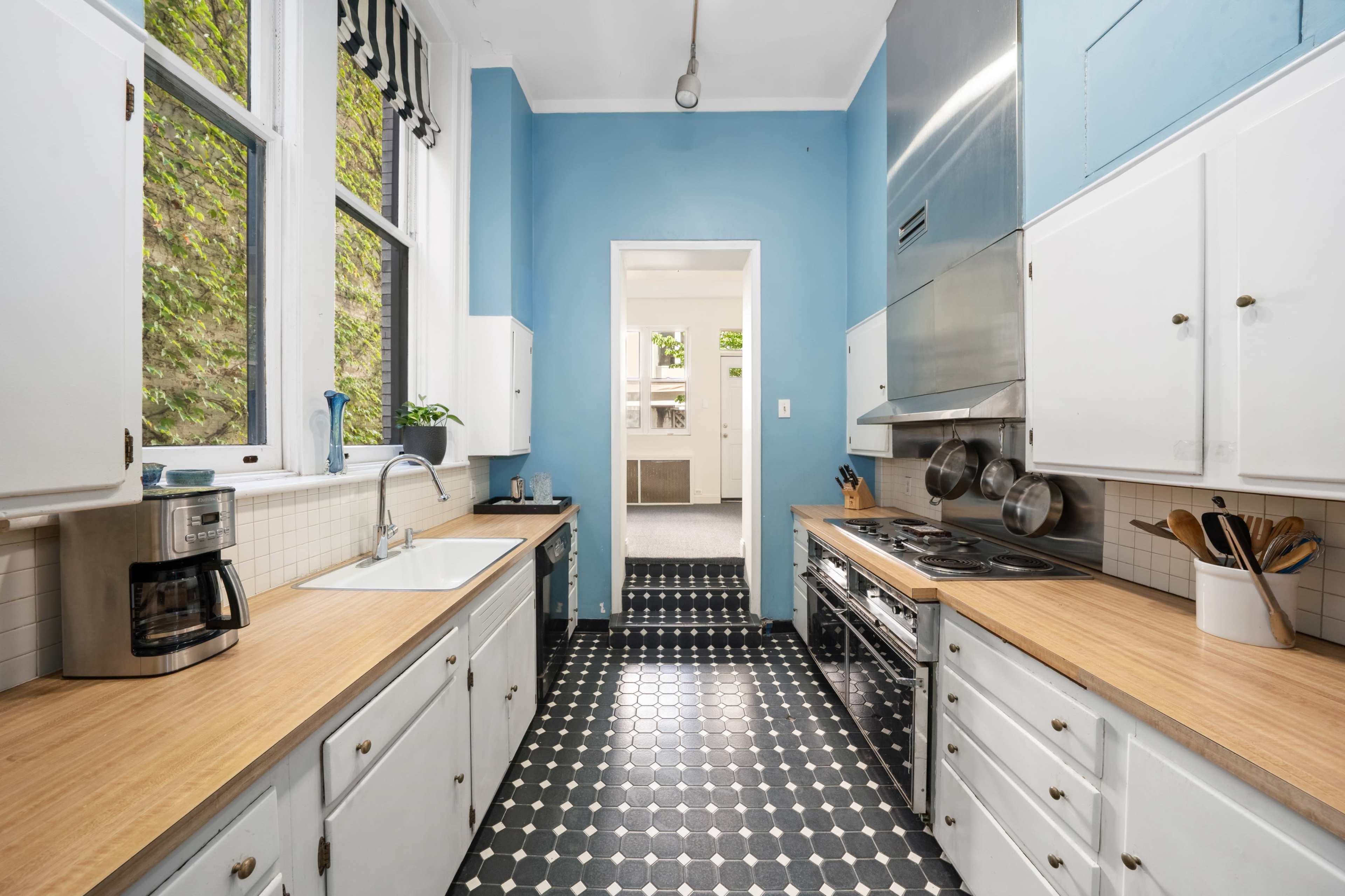 A kitchen with blue walls, a wooden countertop, and black and white tiled flooring, featuring stainless steel appliances and a window allowing natural light.