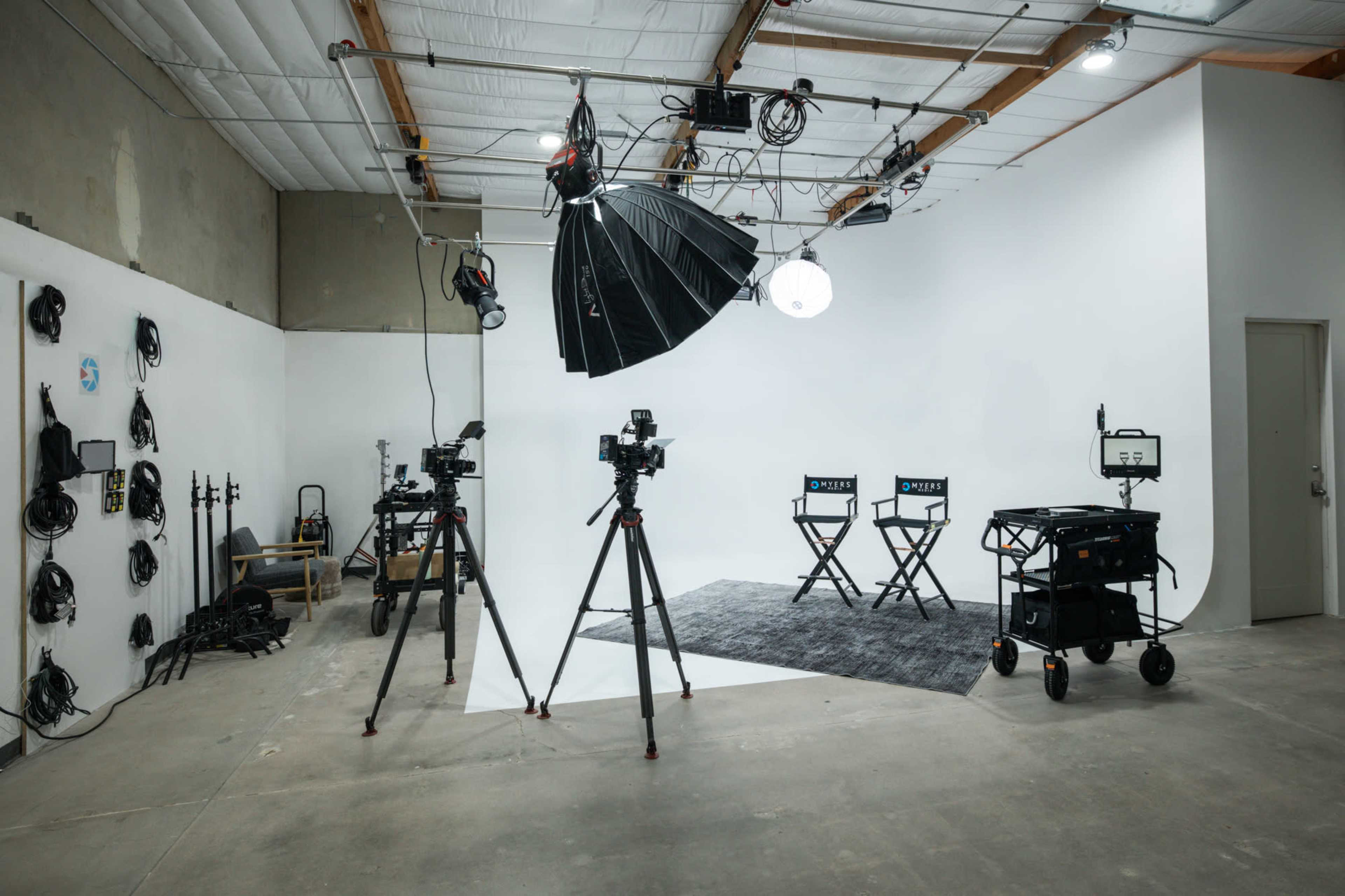 The image shows a photography studio with two director's chairs, lighting equipment, and several cameras set up on tripods against a plain white backdrop.
