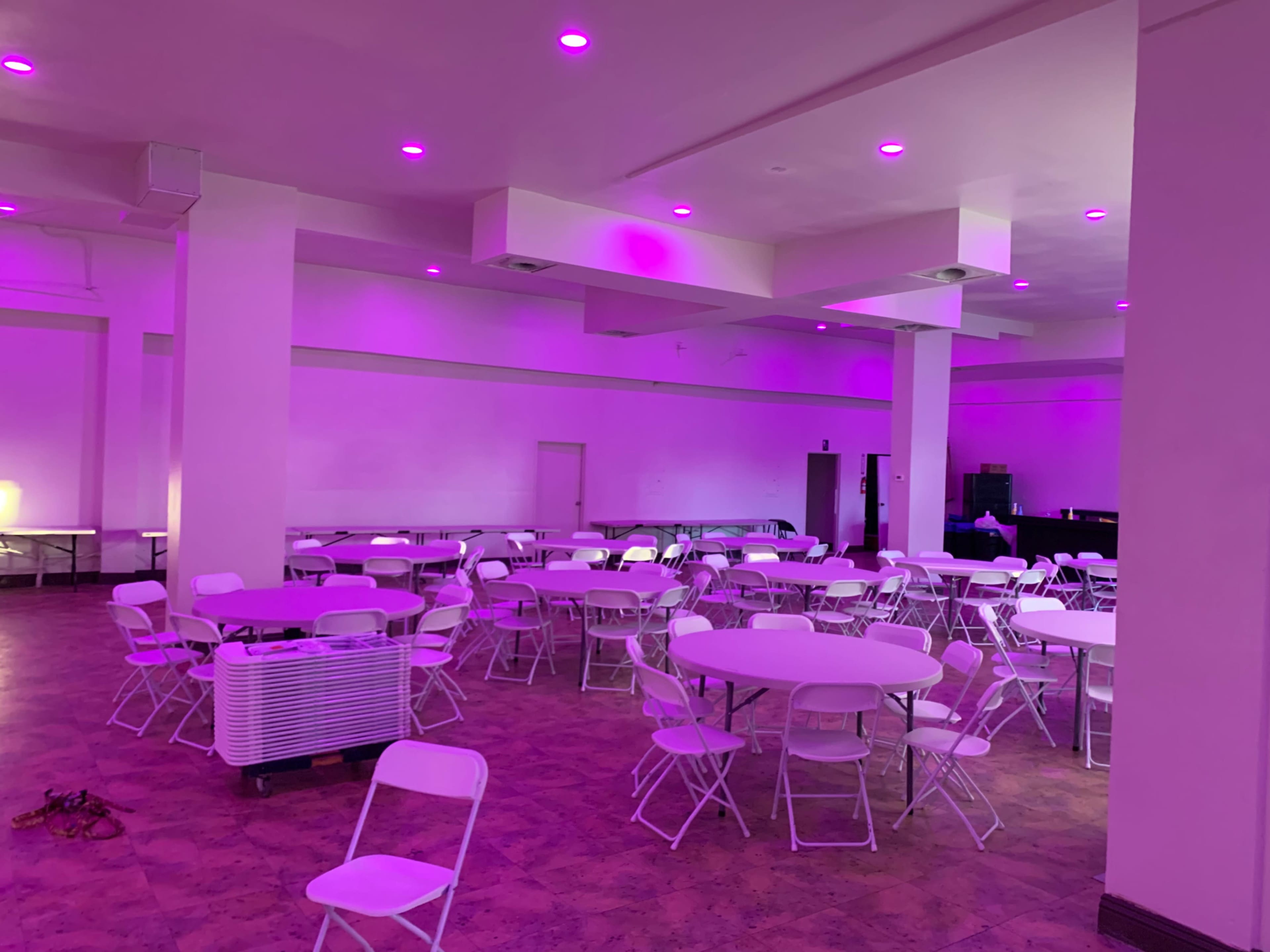 The room features white round tables with folding chairs arranged throughout, illuminated by purple lights on the ceiling.