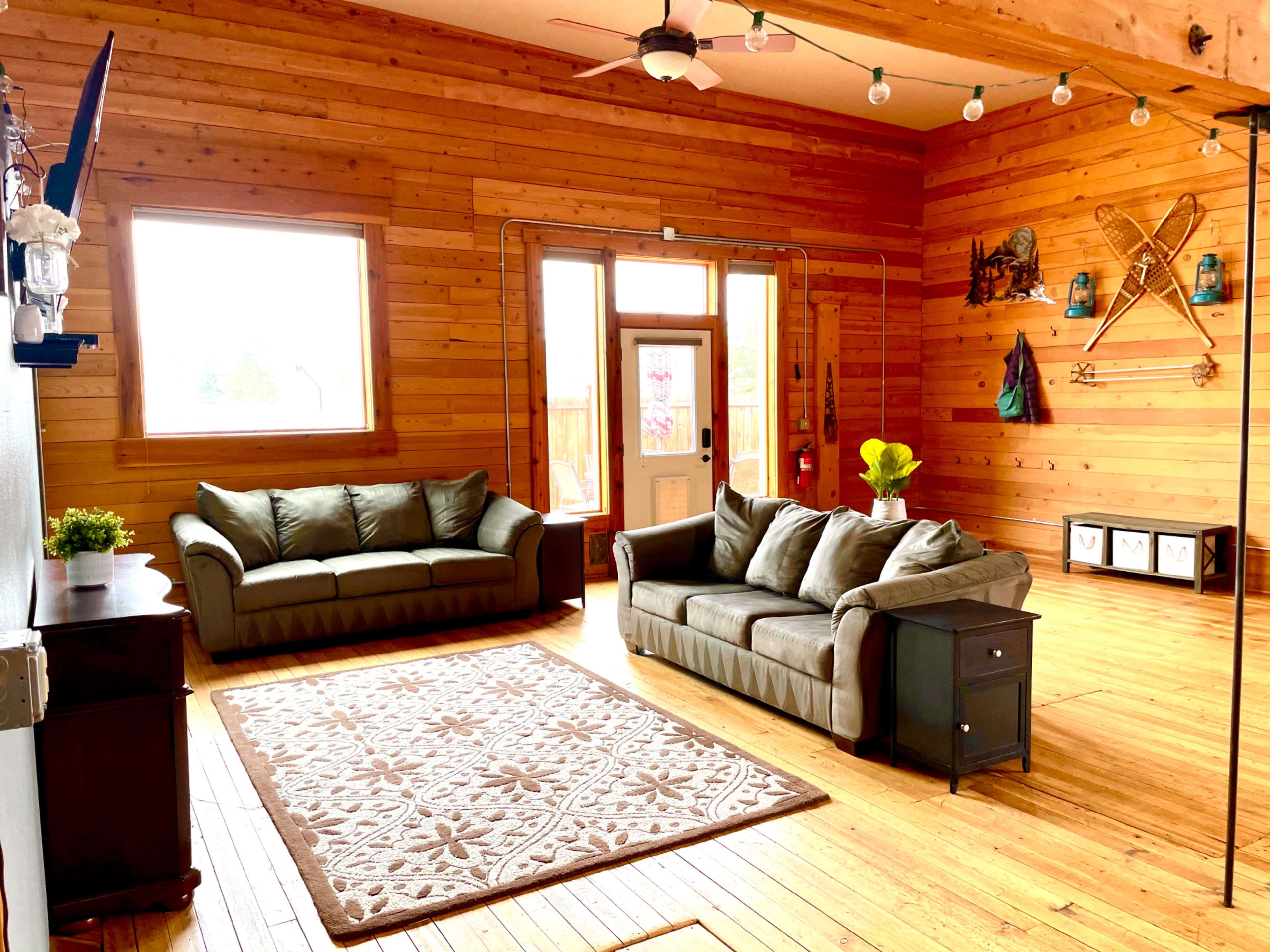 A cozy living room features two green sofas, a decorative rug, and wooden walls, with large windows allowing natural light to enter.