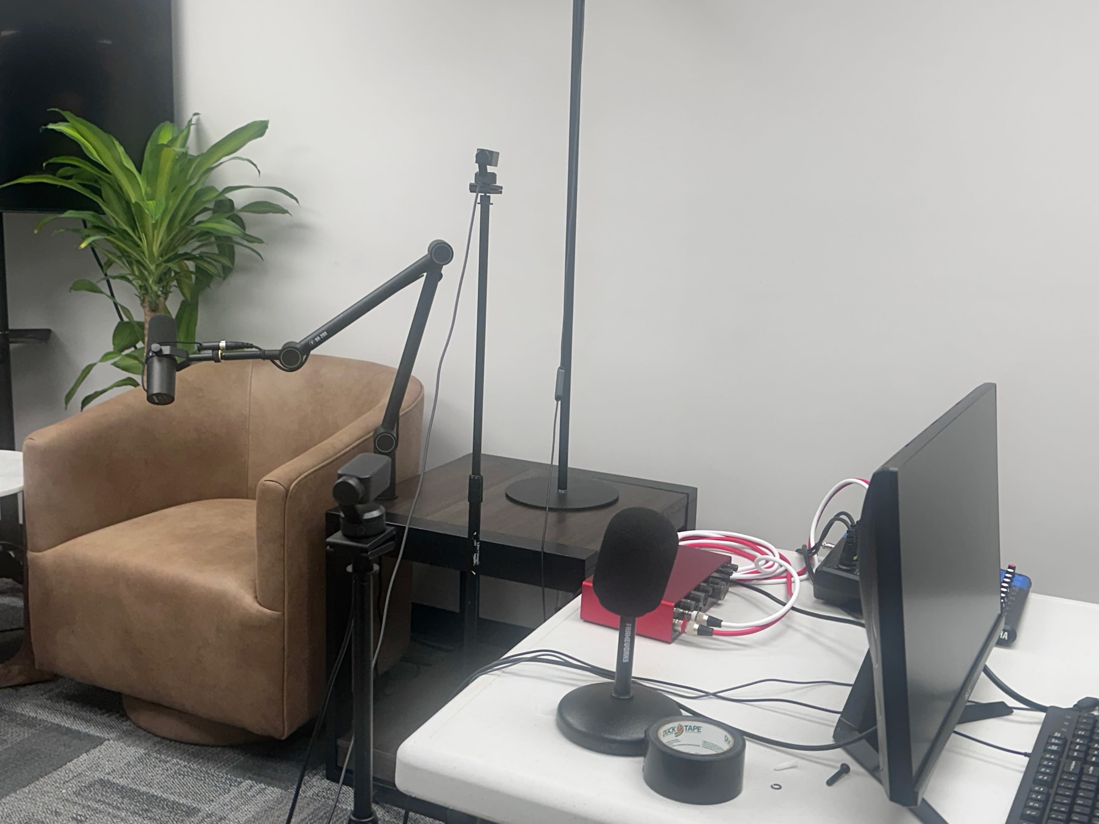 The image shows a small recording setup featuring a microphone on a stand, a computer monitor, and a brown chair against a plain wall with a plant in the background.