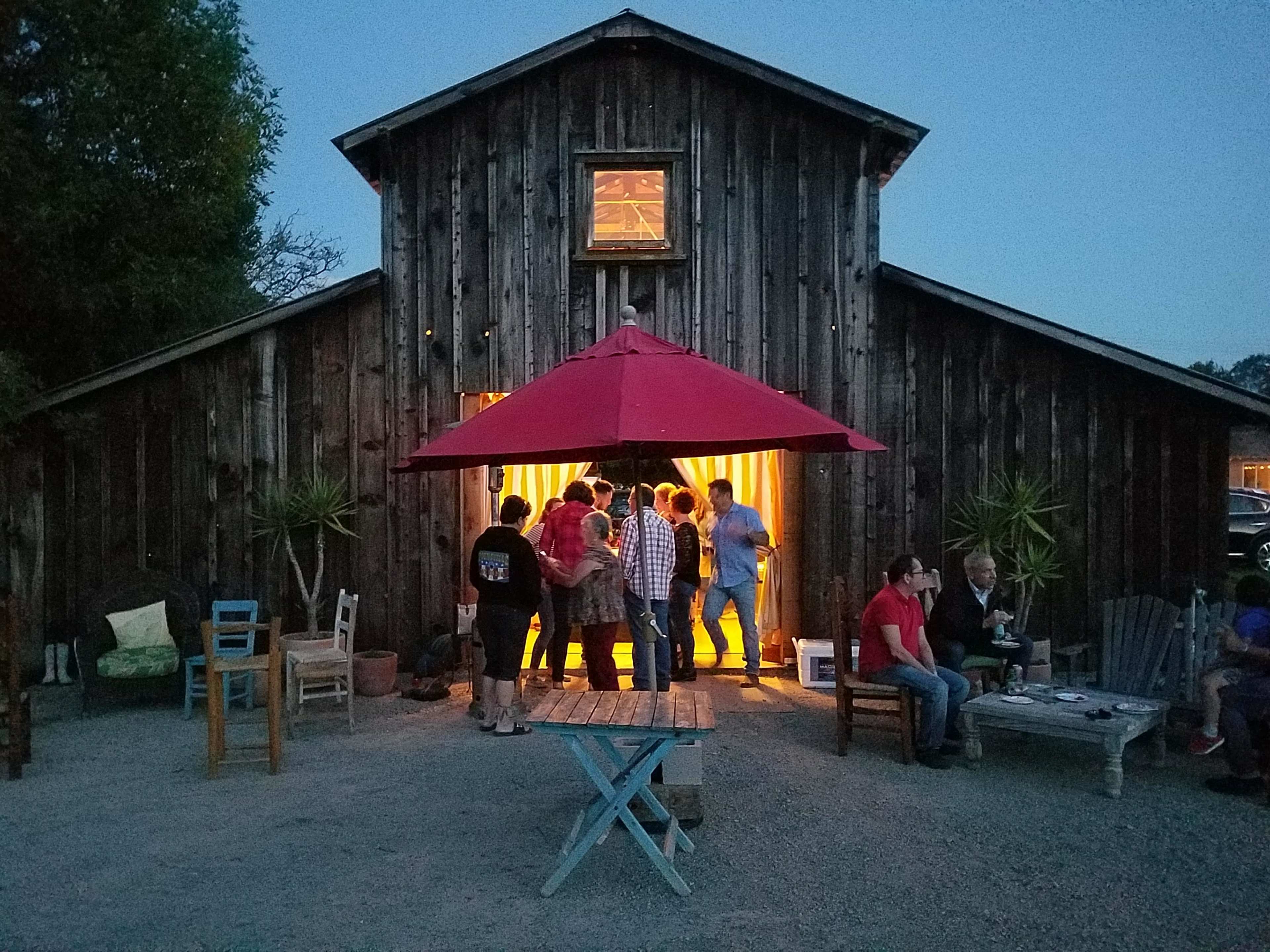 A group of people gathers outside a wooden barn, illuminated from within, as dusk settles around them.