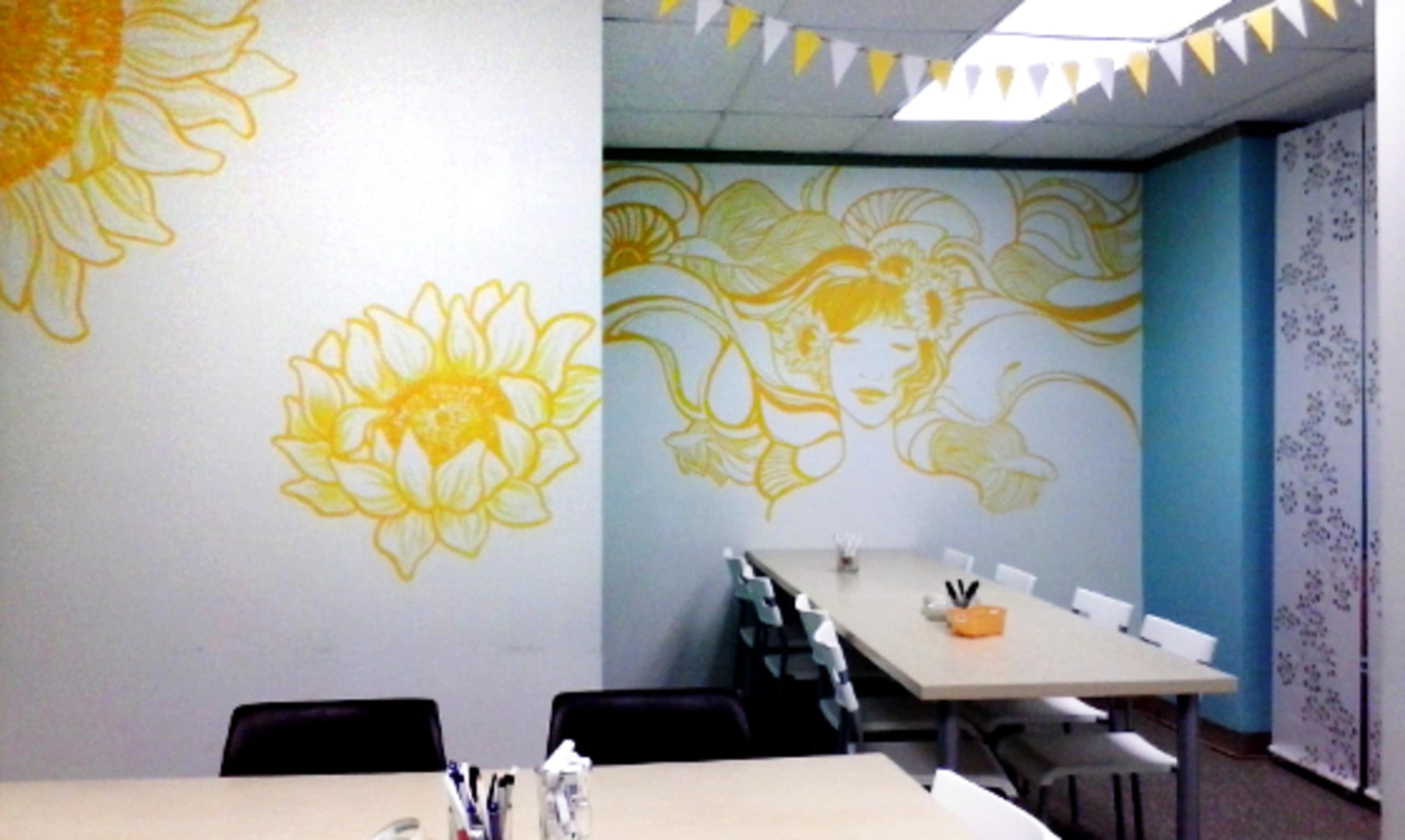 The image shows a brightly decorated office space featuring yellow mural art of sunflowers and a stylized figure on the walls, with a central table and chairs arranged for meetings.
