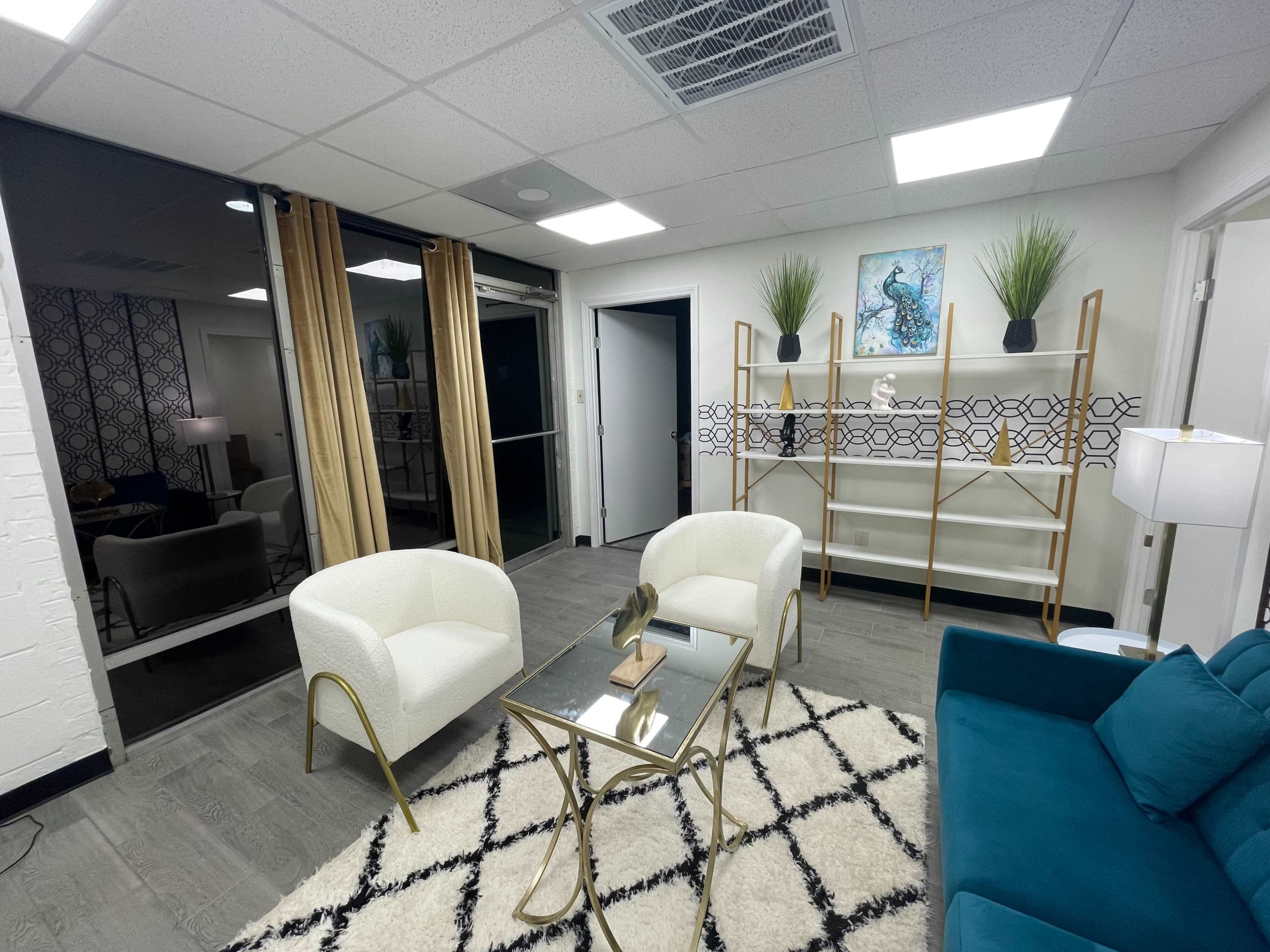 A modern waiting area with two white chairs, a teal sofa, a glass coffee table, and a decorative shelving unit against a patterned wall.
