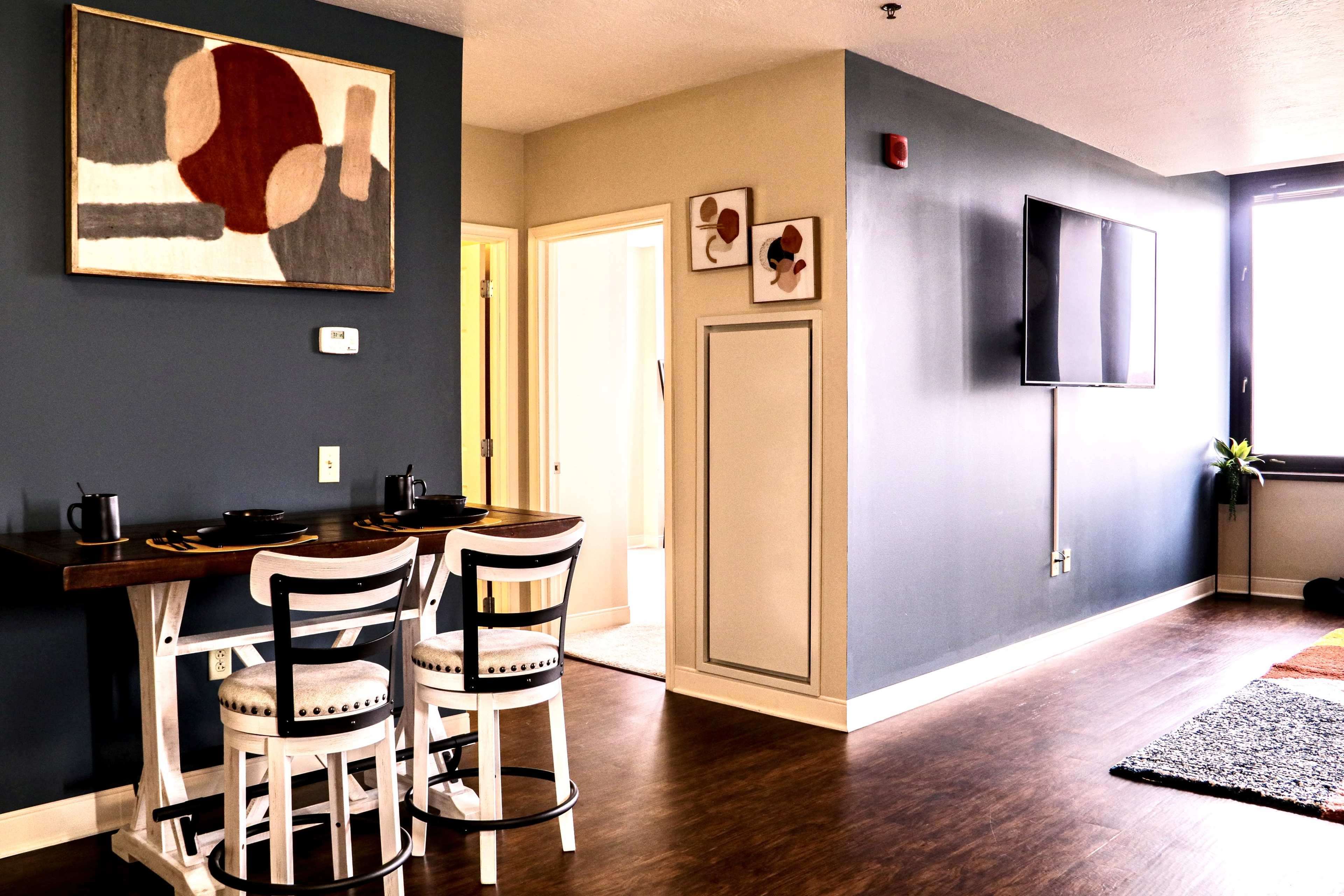 The image shows a modern living space with a dining area featuring two stools, a wall-mounted TV, artwork, and a doorway leading to a bathroom.