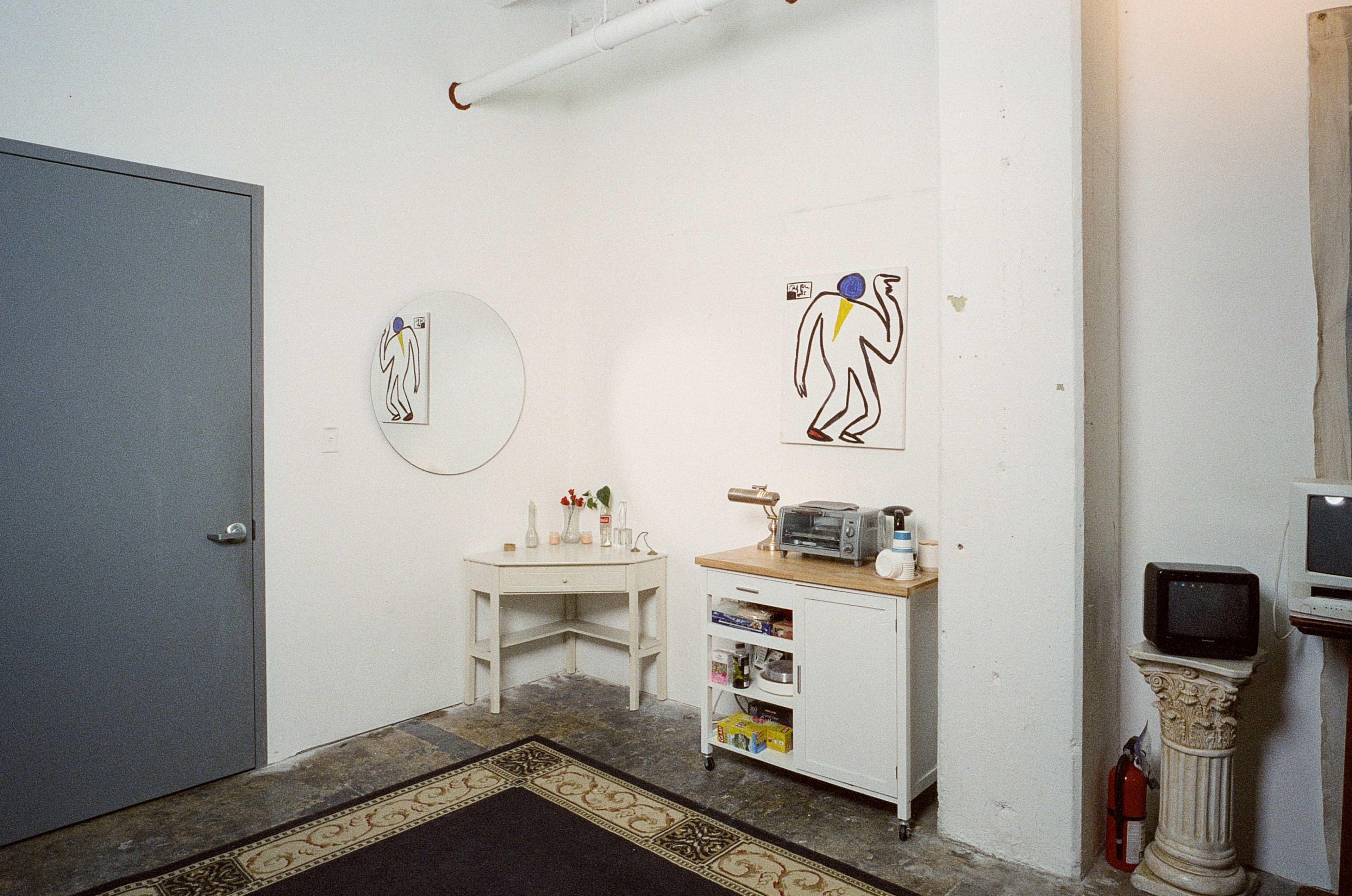 A corner of a minimalist room featuring a gray door, a round mirror, a small white table with flowers, a cart with kitchen items, and an old television near a column.