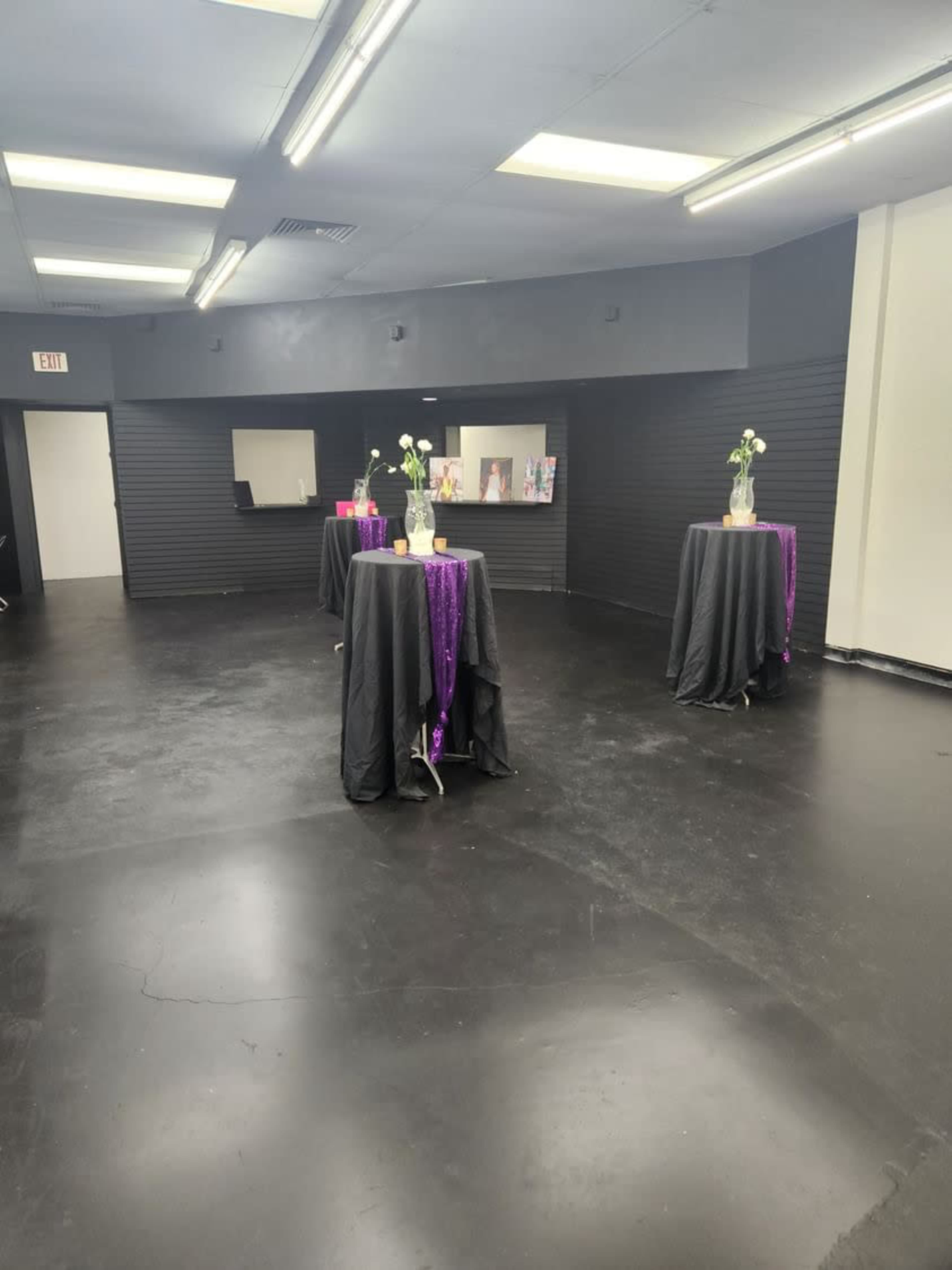 A spacious, dimly lit room with three tall tables covered in black cloth, each adorned with a vase of white flowers and purple table runners.