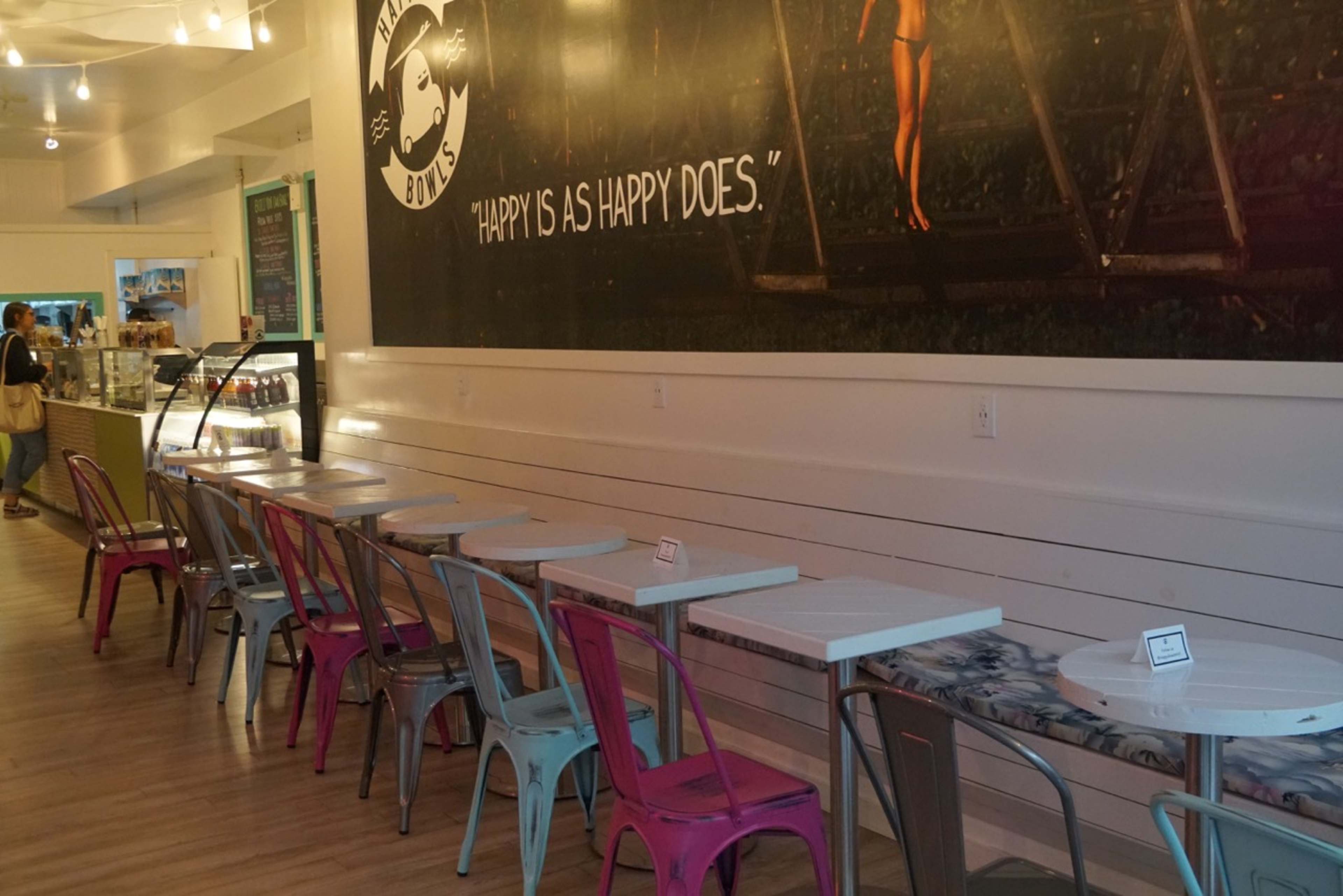 A row of tables with colorful metal chairs is set up in a café, with a large wall mural featuring a person and the quote "HAPPY IS AS HAPPY DOES."