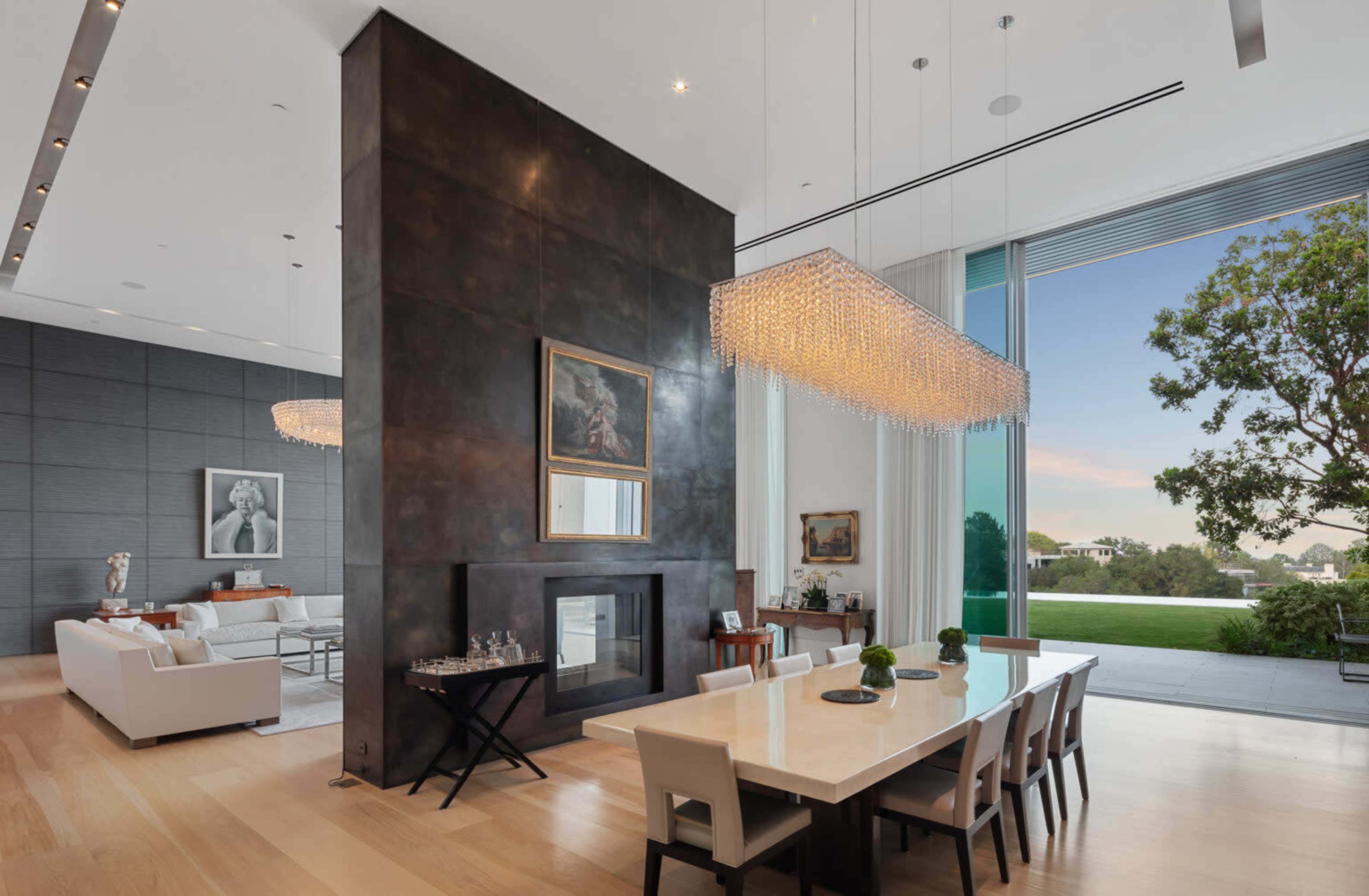 A modern living space featuring a large dining table, a contemporary chandelier, and a wall-mounted fireplace, with floor-to-ceiling windows overlooking a green landscape.