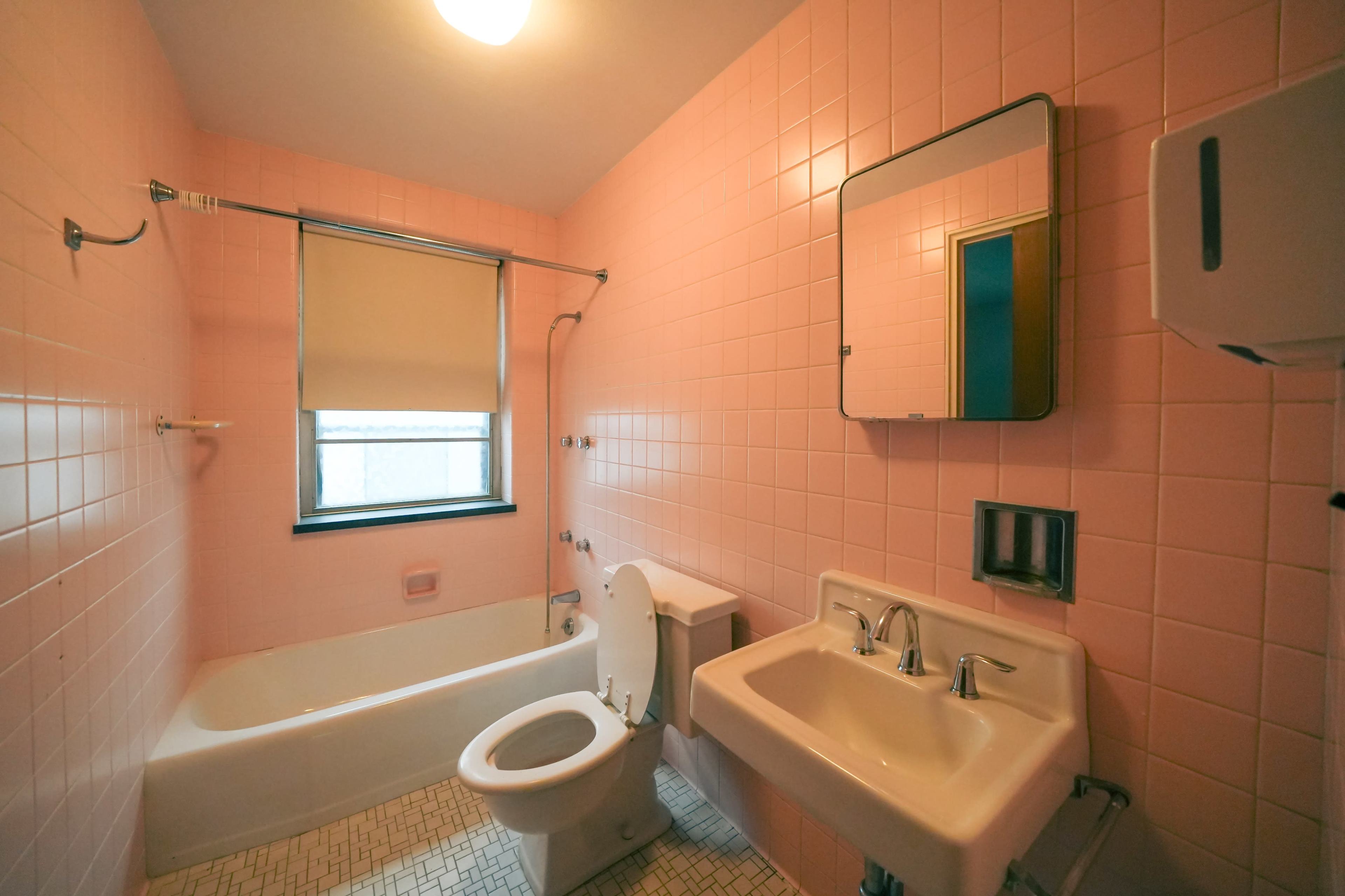 A bathroom with pink tiled walls, a bathtub, toilet, and a sink, featuring a window with a roller shade.