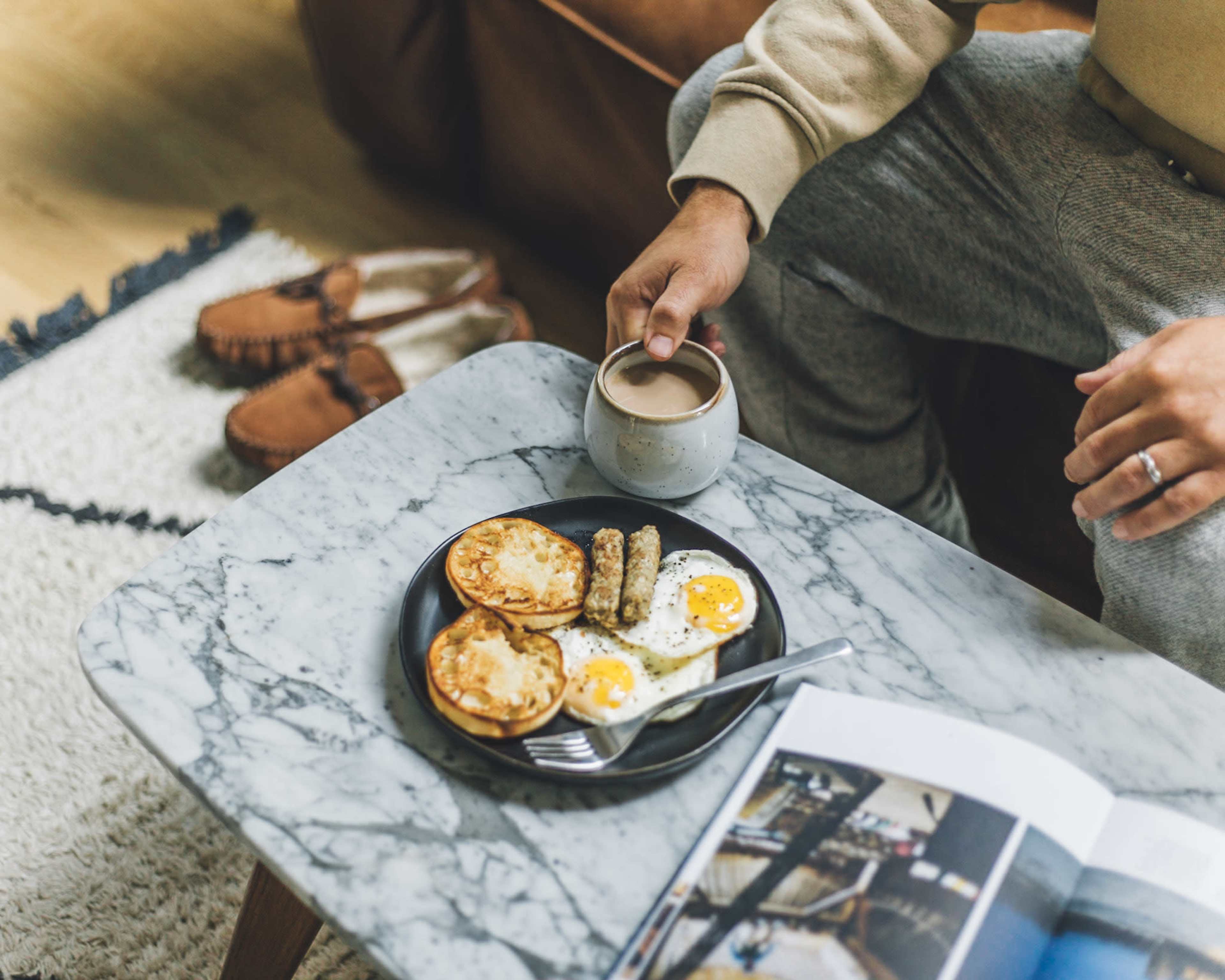 A man sits on a couch, holding a cup of coffee, with a plate of fried eggs and sausage links on a marble coffee table beside a magazine and slippers.