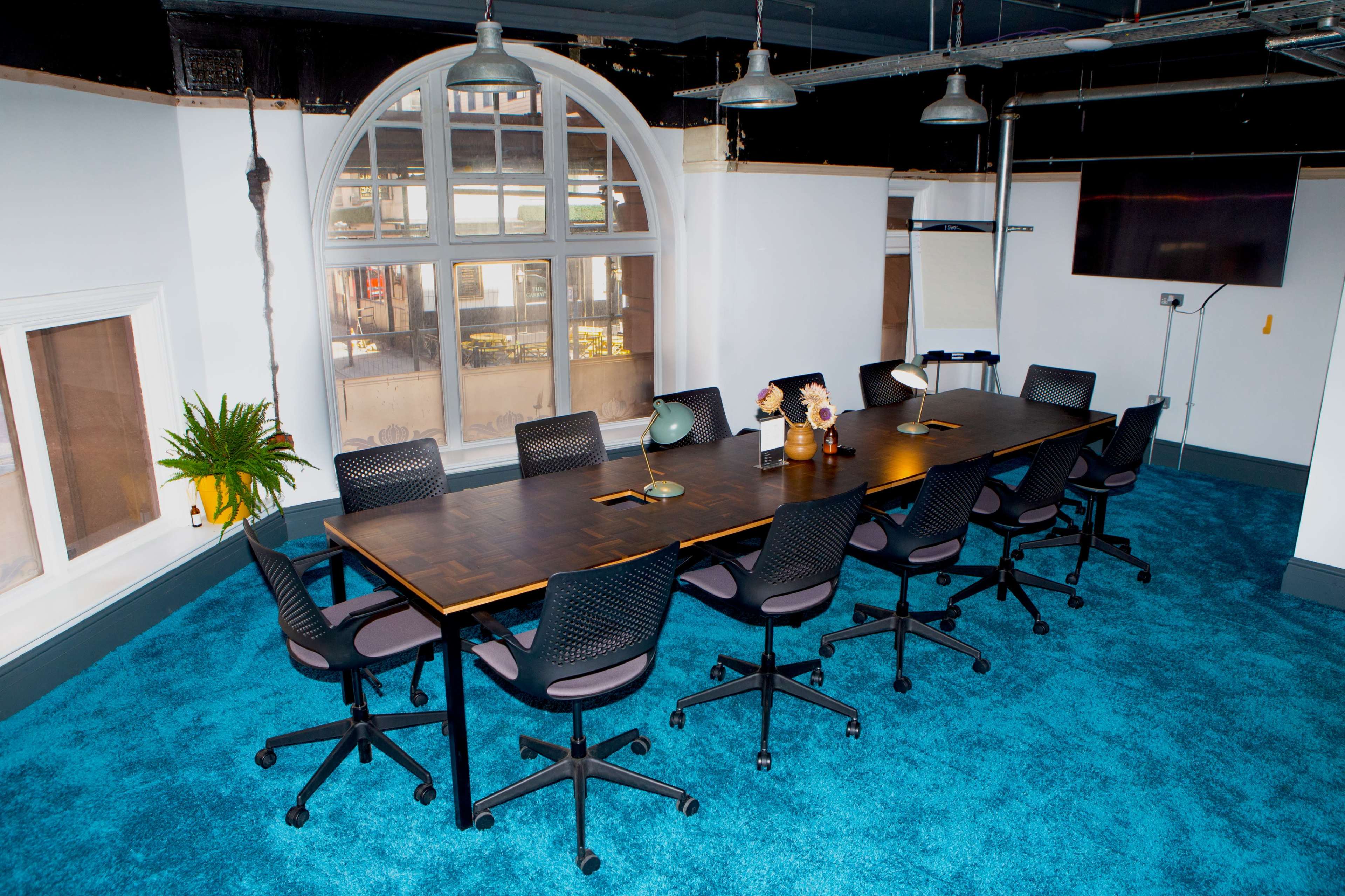 A long conference table with eight chairs is centered in a brightly lit meeting room featuring a large arched window and blue carpet.