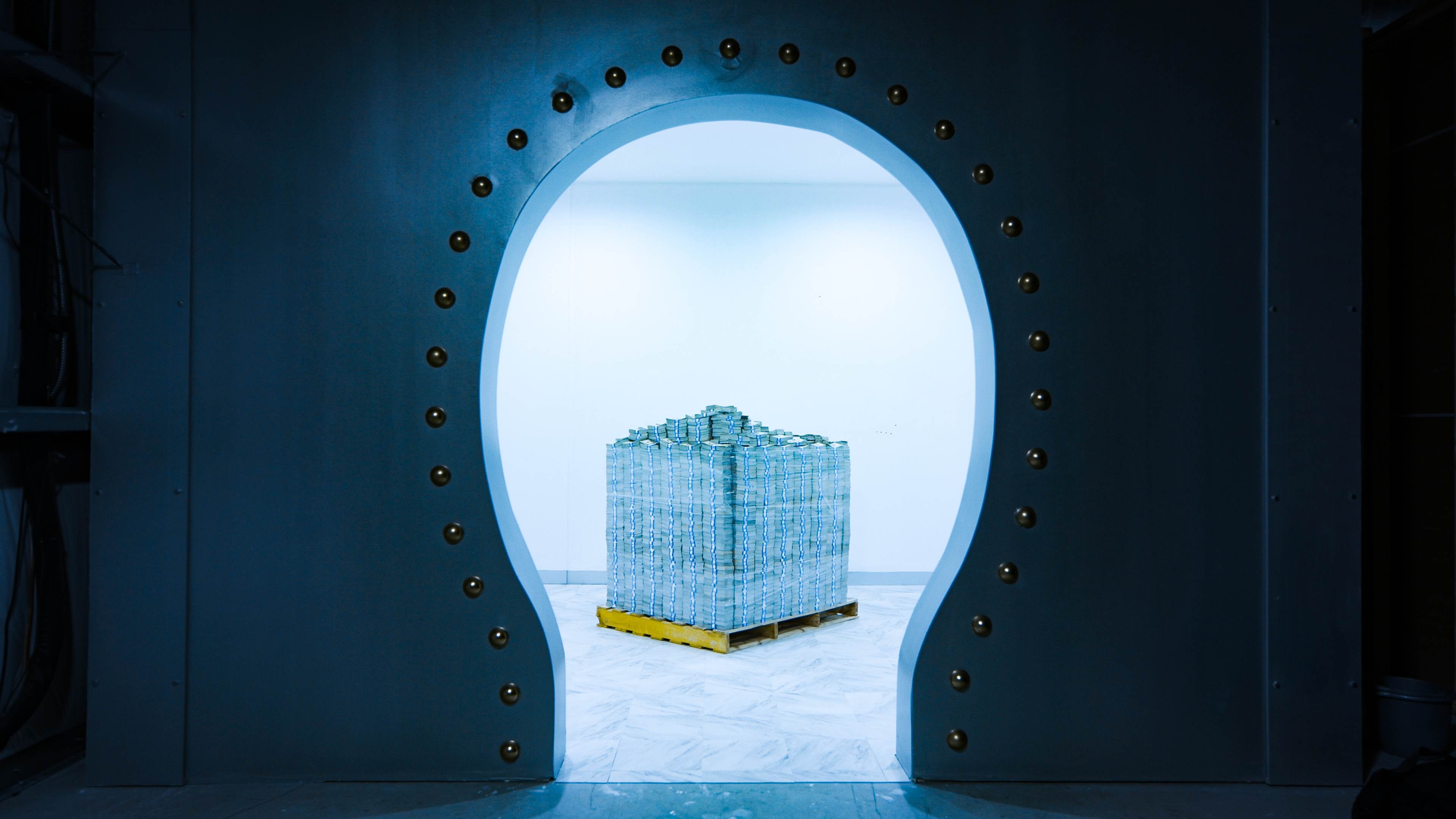 A stack of cash is visible behind a large, round doorway in a well-lit room.