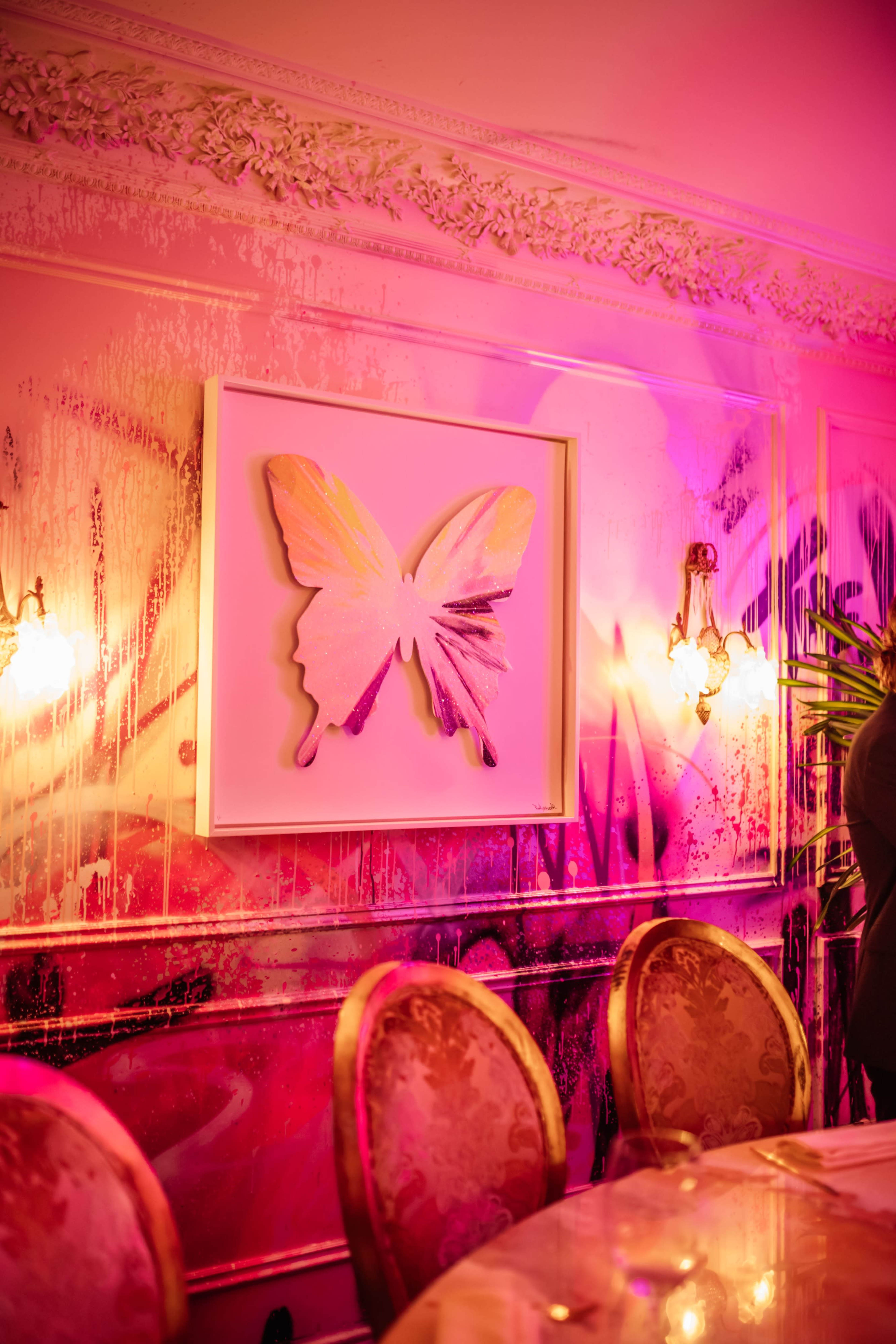 A butterfly artwork is framed on a brightly lit wall with colorful splashes and ornate sconces casting warm light in a dining area.