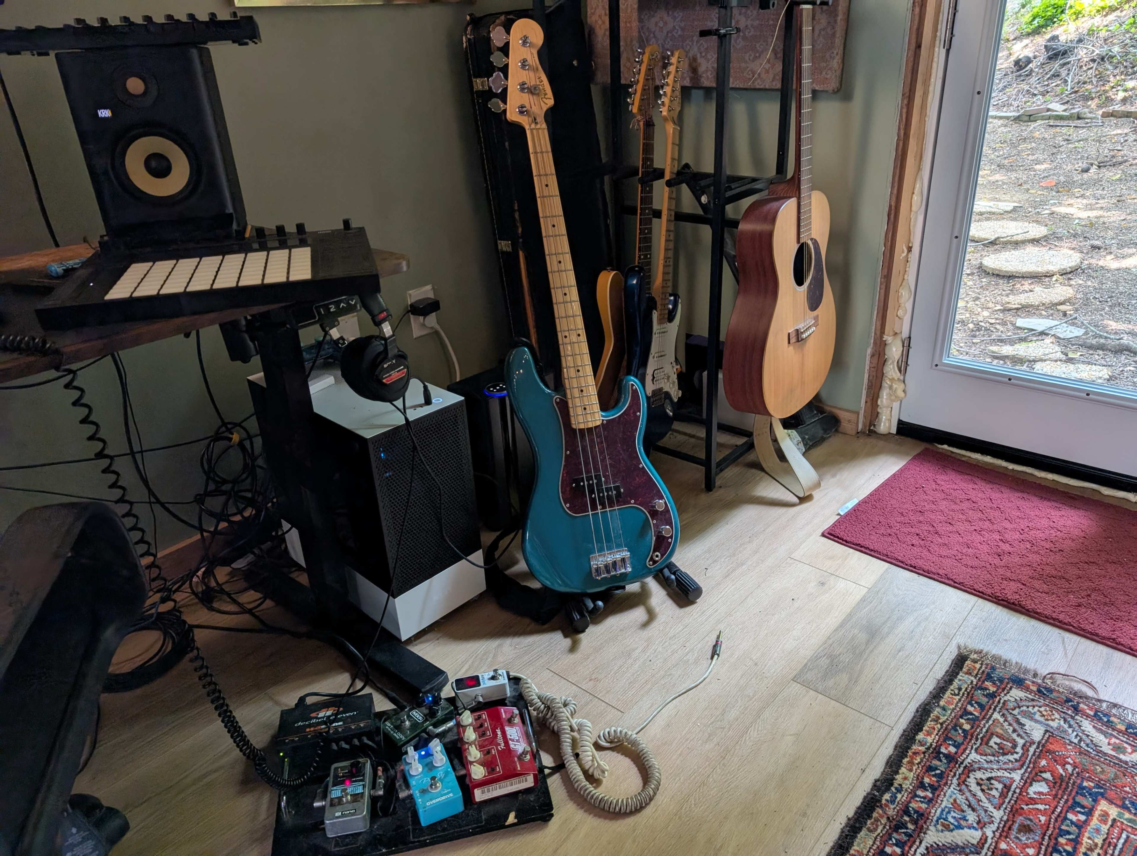 A blue electric bass guitar rests on the floor beside a stack of music equipment and an acoustic guitar near an open door.