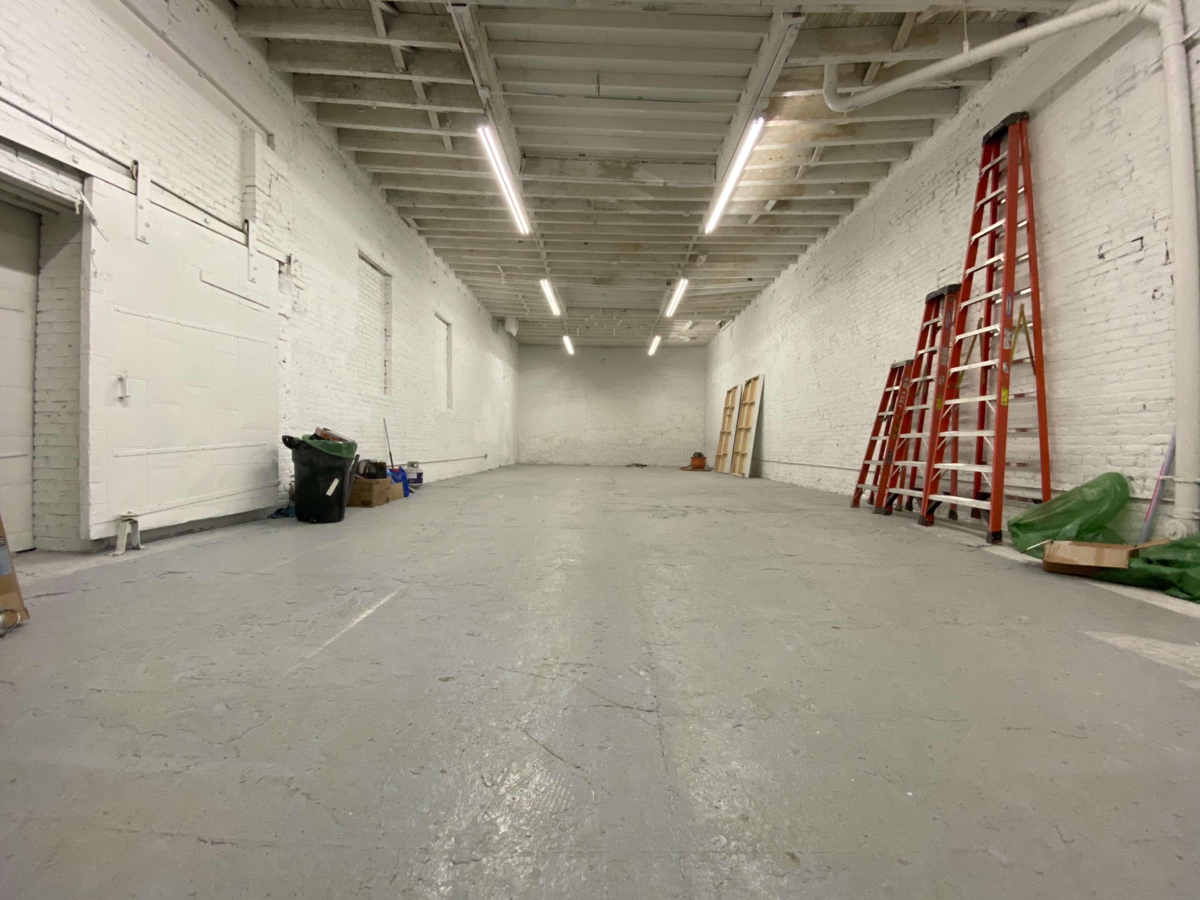 The image shows a large, empty warehouse space with white brick walls, a polished gray floor, a ladder against the wall, and various items scattered around.