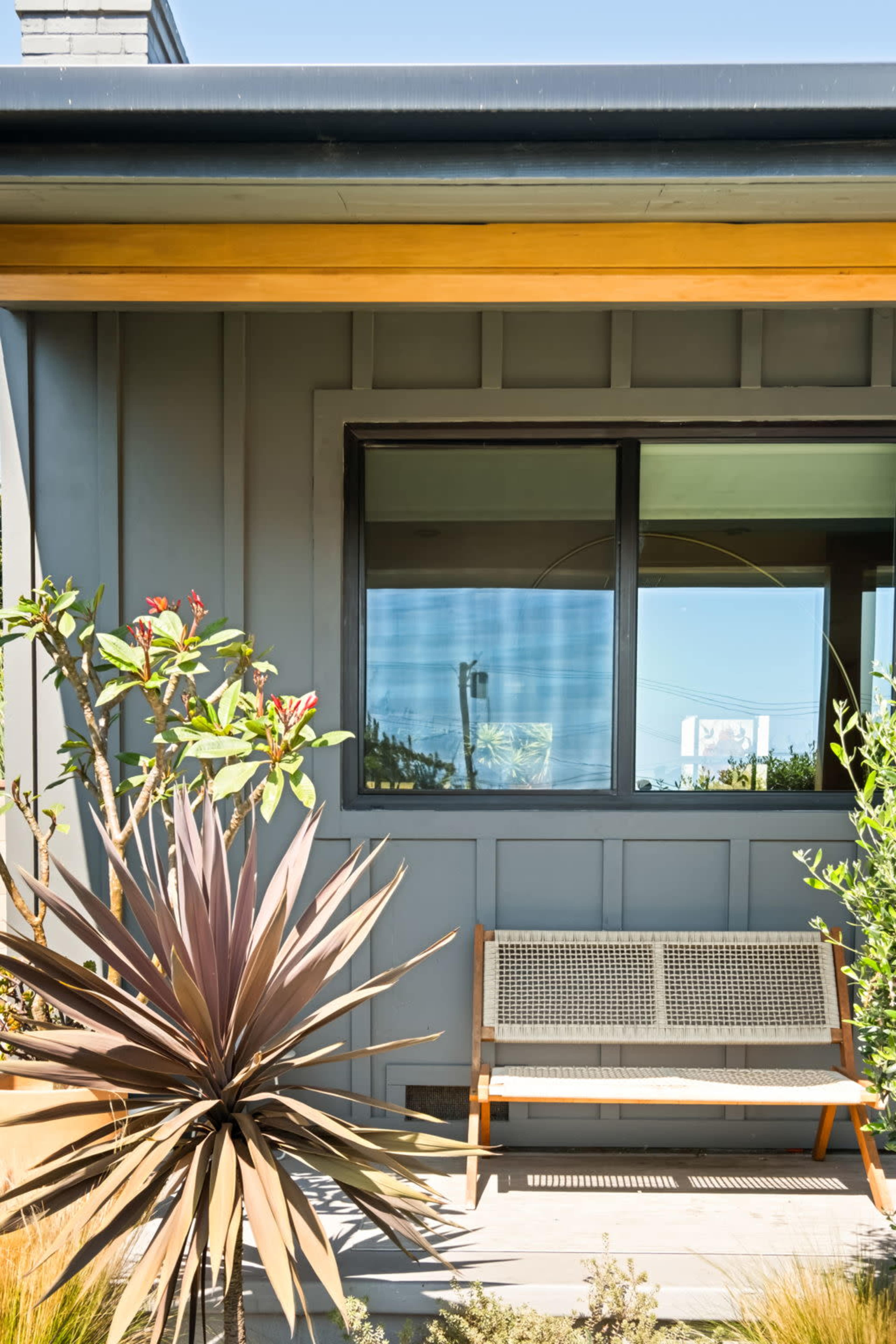 A modern house features a gray exterior, a large window, and a bench surrounded by plants.