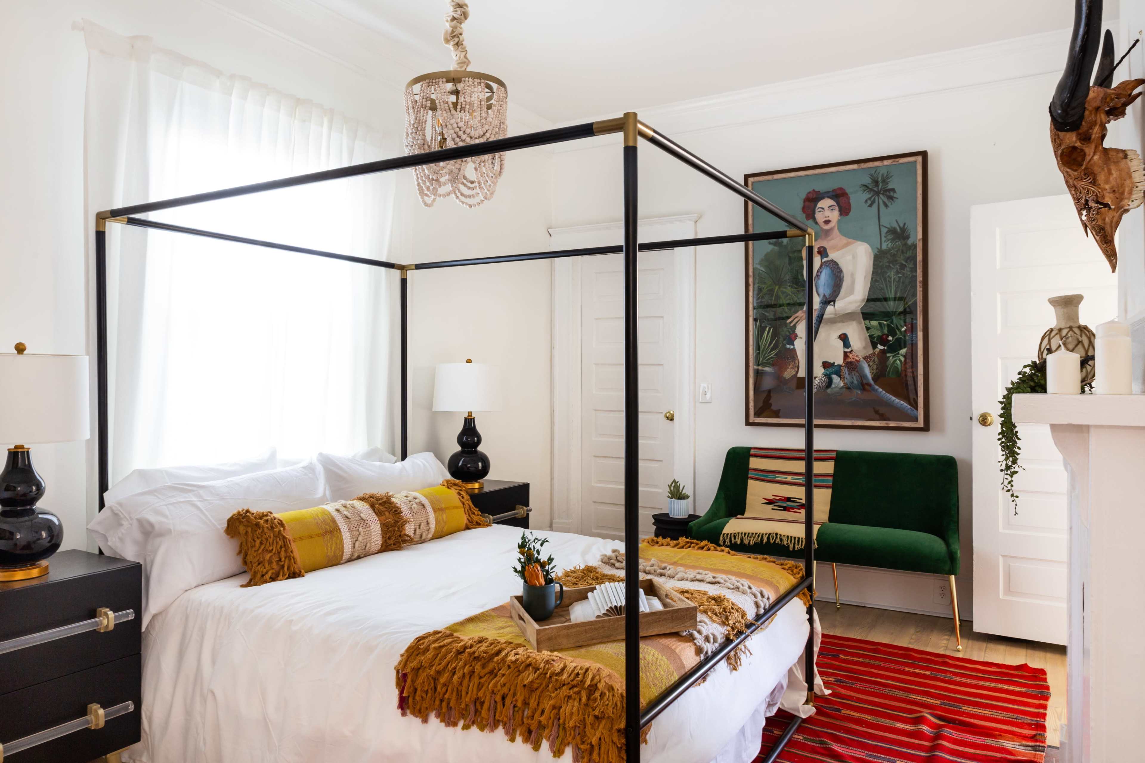 A contemporary bedroom features a four-poster bed with a white canopy, colorful throw blankets, a green settee, and an oversized artwork on the wall.