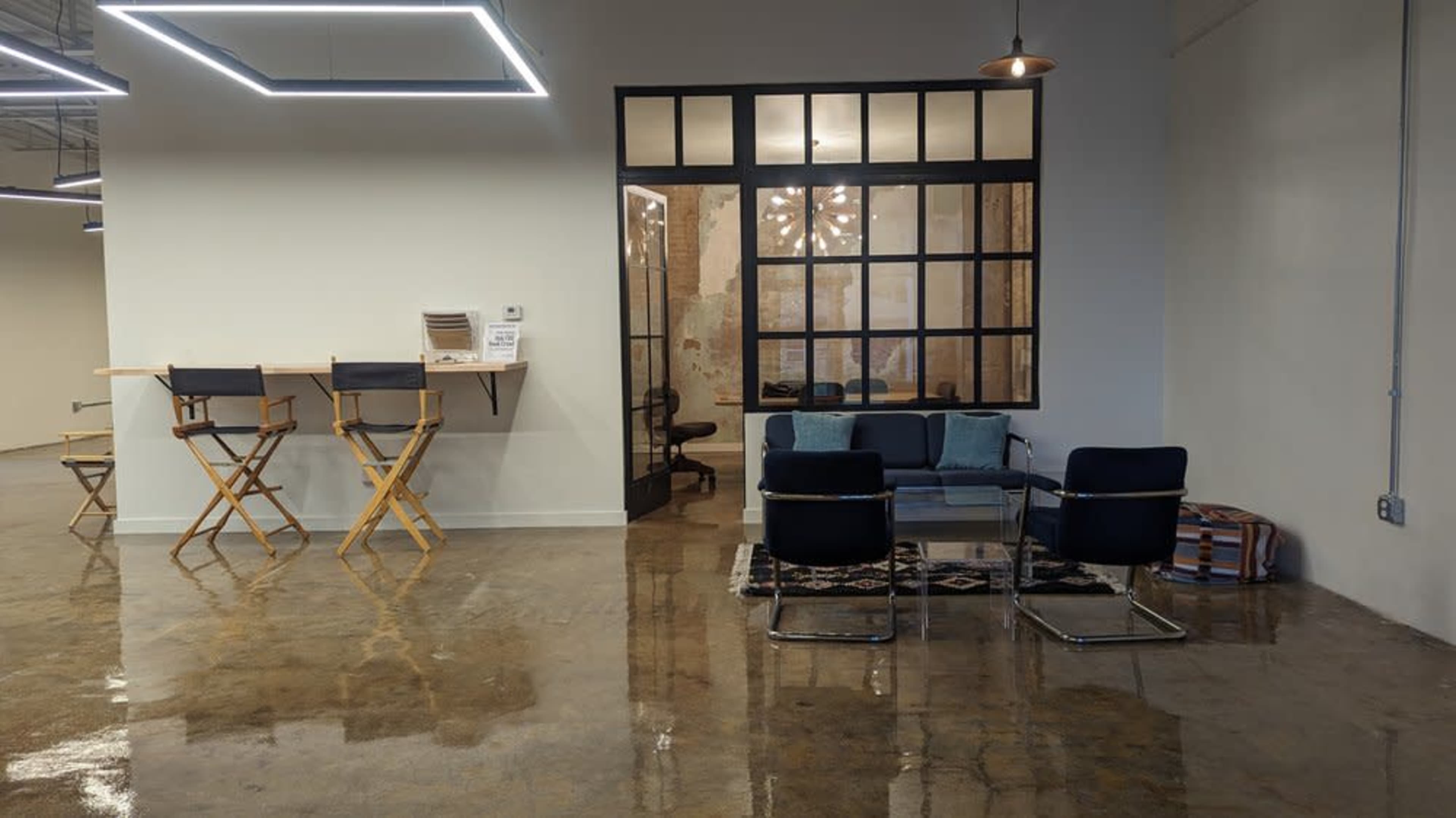 The image shows a modern office space featuring a seating area with a blue couch and glass coffee table, along with a small bar-like counter and director's chairs against one wall.
