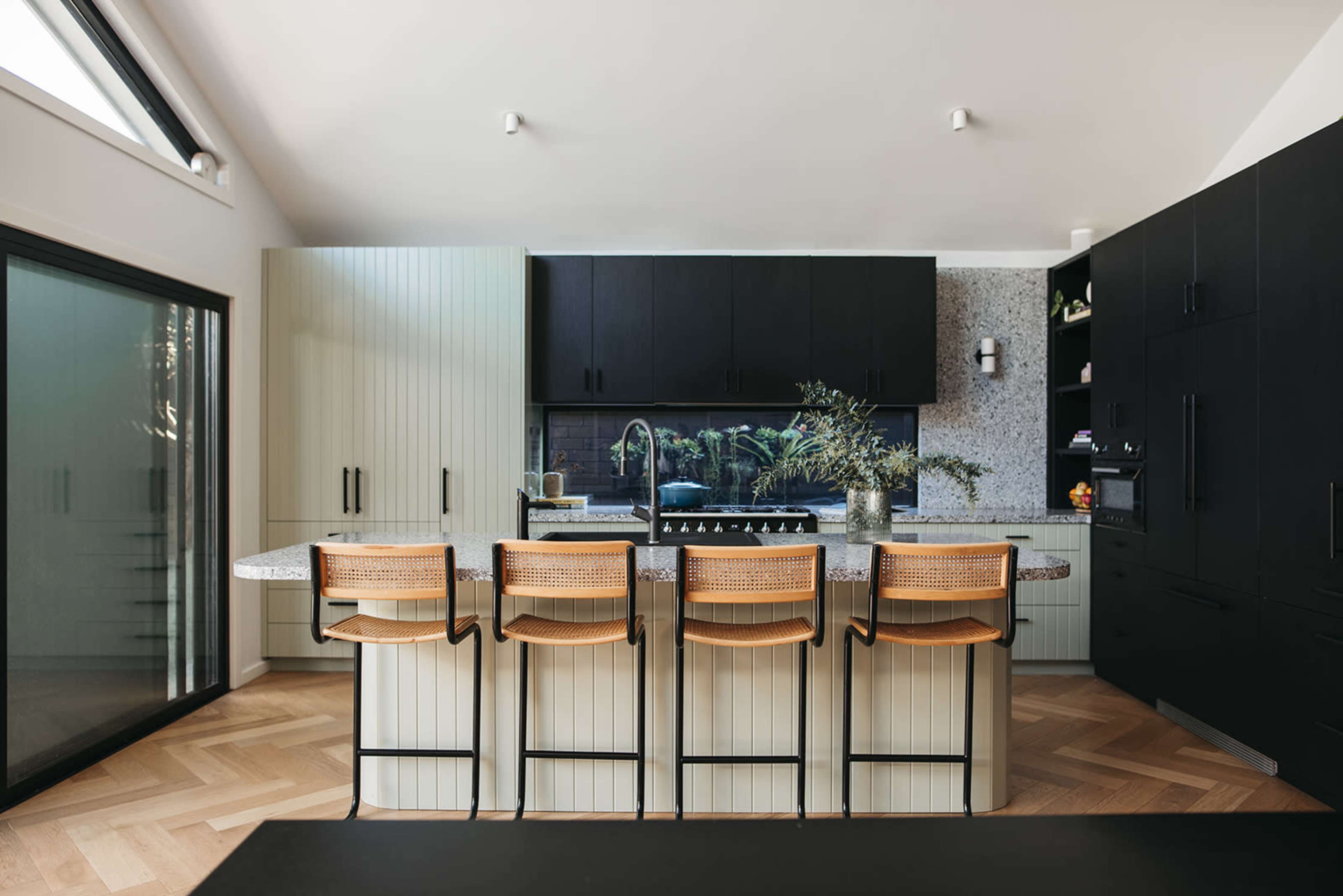 A modern kitchen features a large island with four woven bar stools, black cabinetry, and a light green pantry against a textured wall.