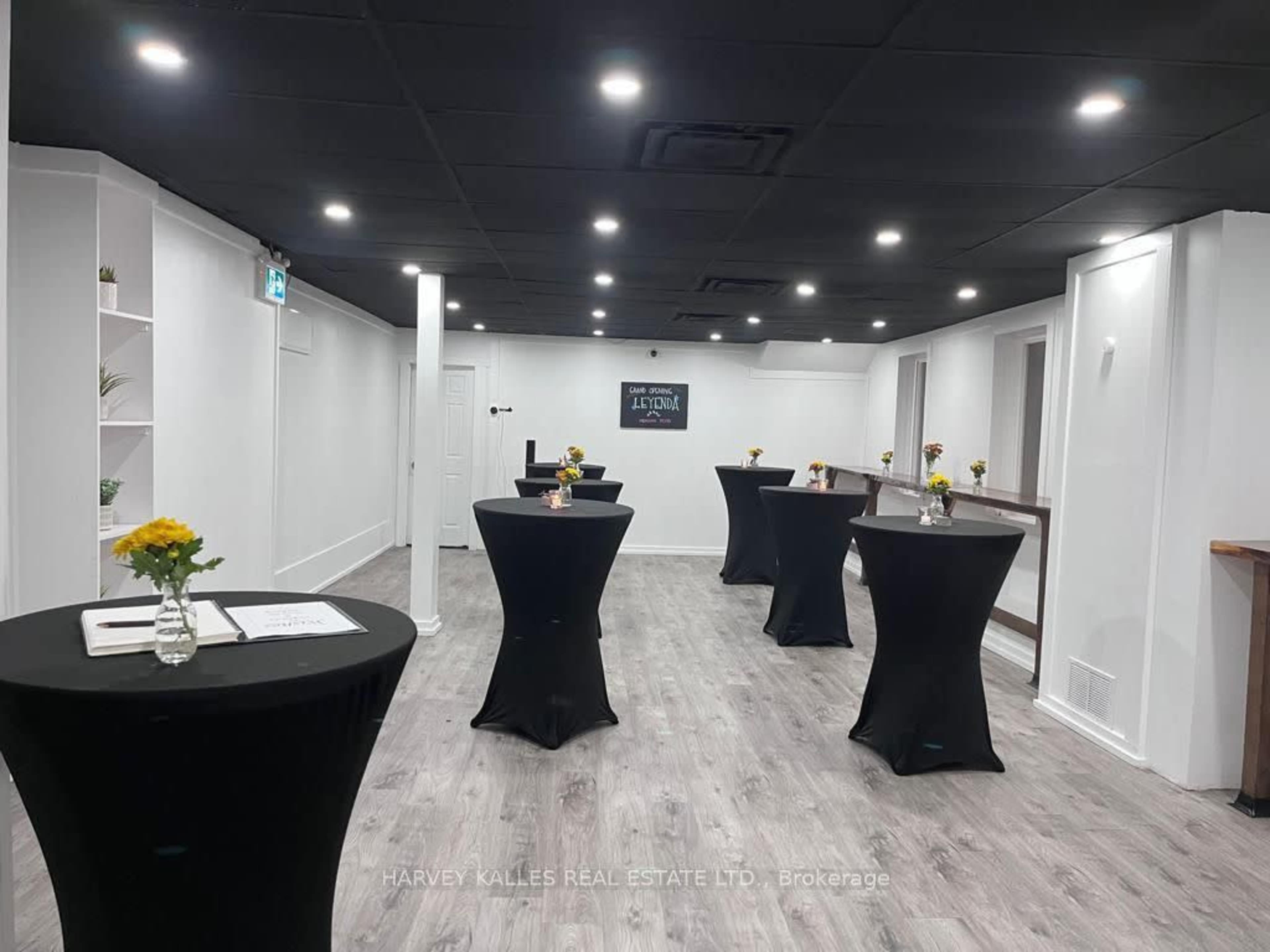 The image depicts a modern event space with black cocktail tables adorned with small flower arrangements and bright lighting.