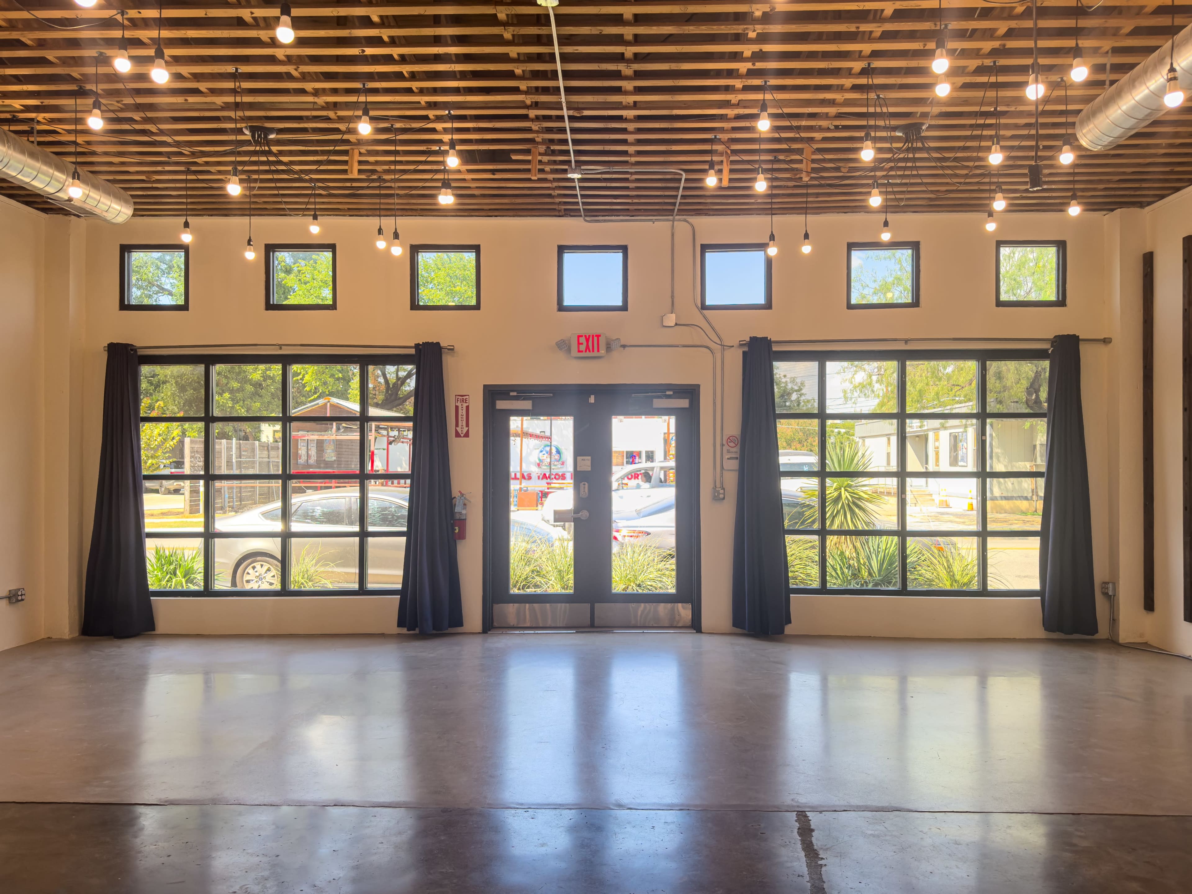 The image shows a spacious interior room with large windows, a double door entrance, and hanging lights, featuring a polished concrete floor and curtain-covered windows.