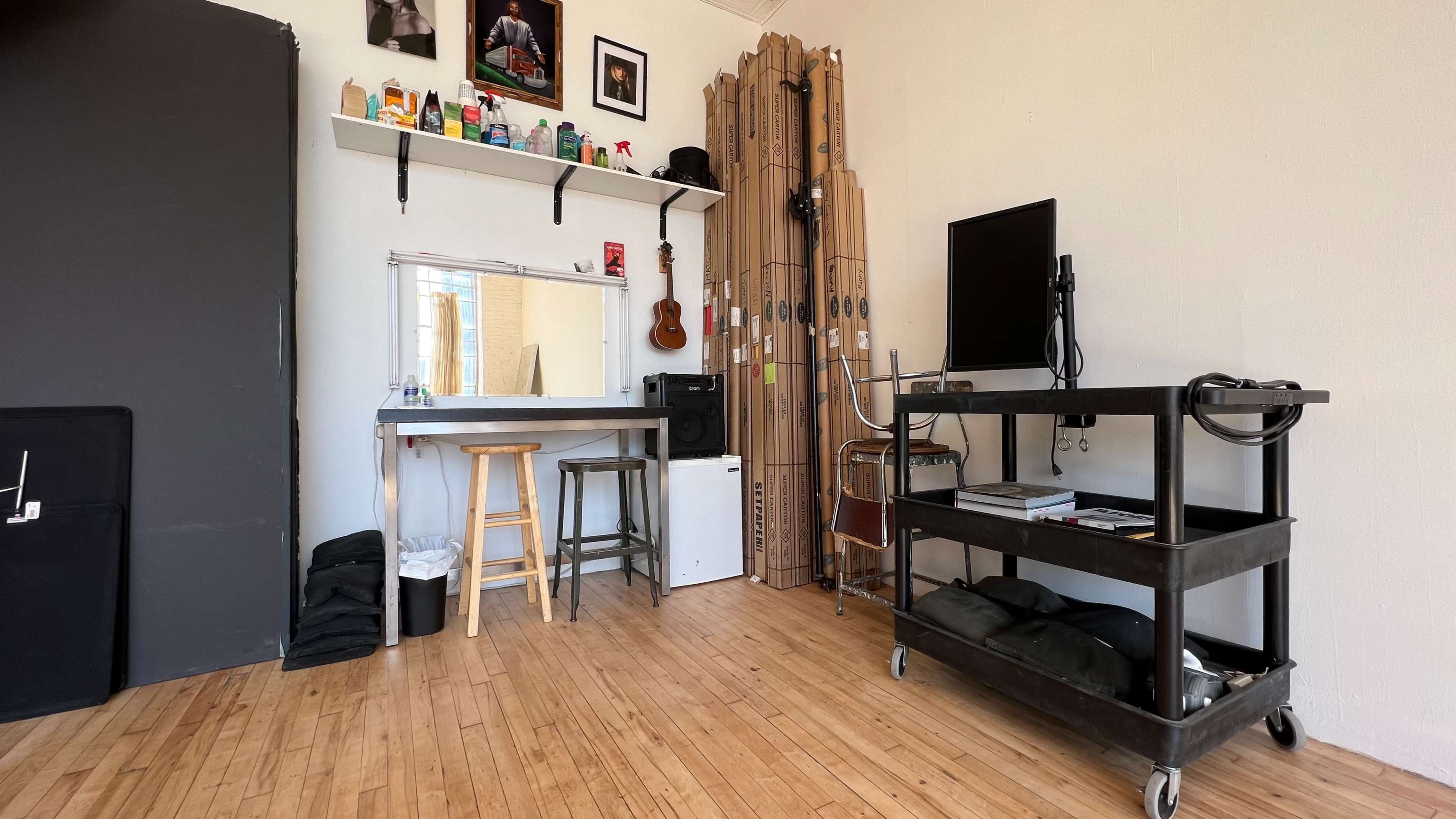 The image shows a workspace with a mirror, a stool, a shelf filled with various items, a television on a cart, and stacked cardboard tubes against the wall.